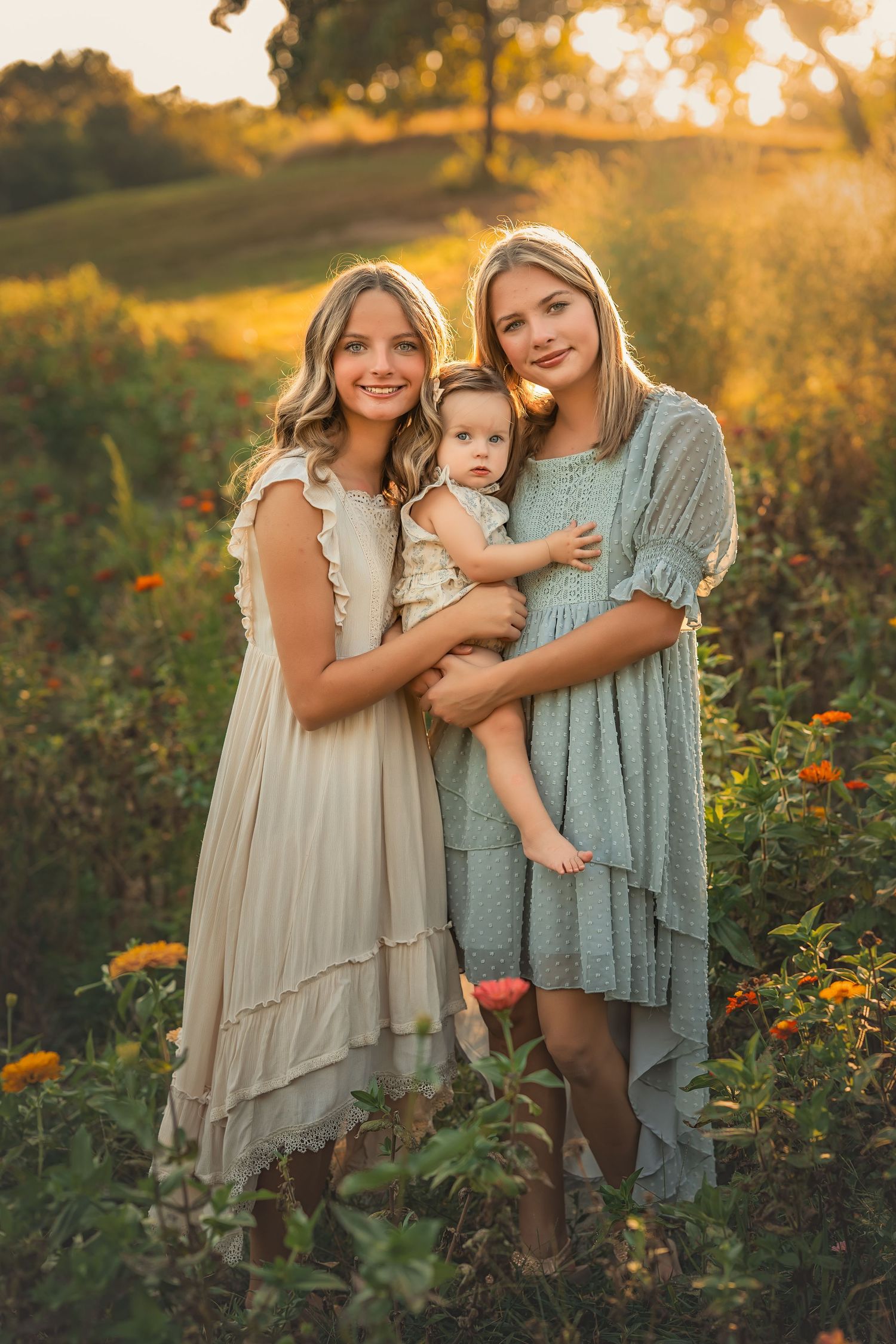 Two teenage girls in flowy dresses hold a baby girl in a sunlit flower field at golden hour.
