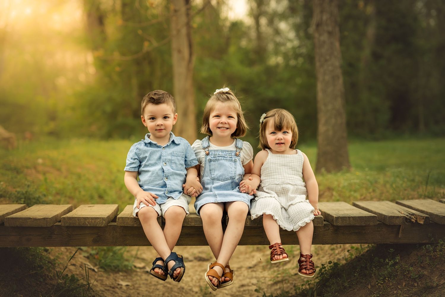 Three young children sitting together on a wooden bench in a sunlit park, smiling and holding hands.