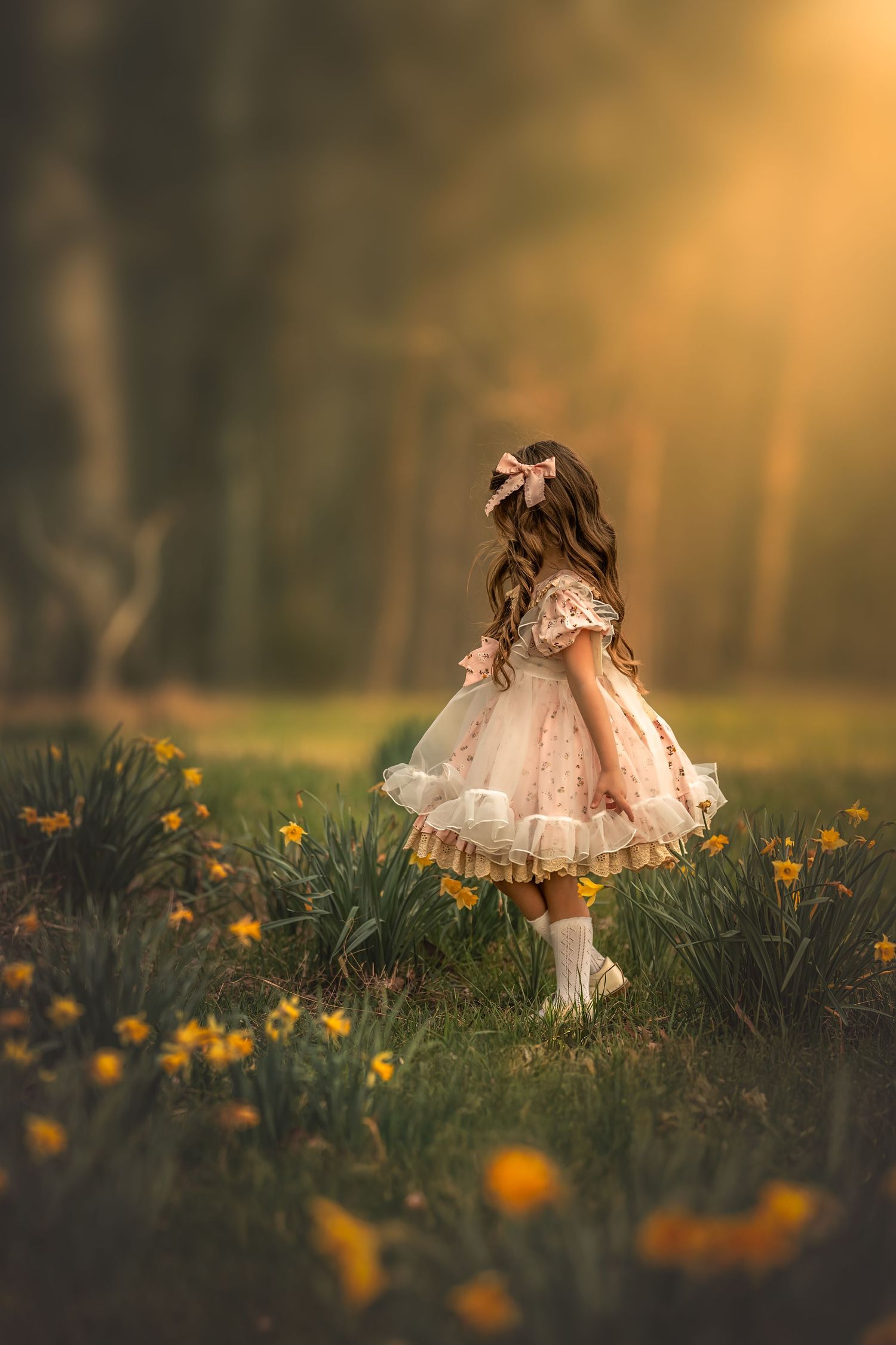 A young girl in a vintage pink dress walks through a field of yellow daffodils in golden sunset light.