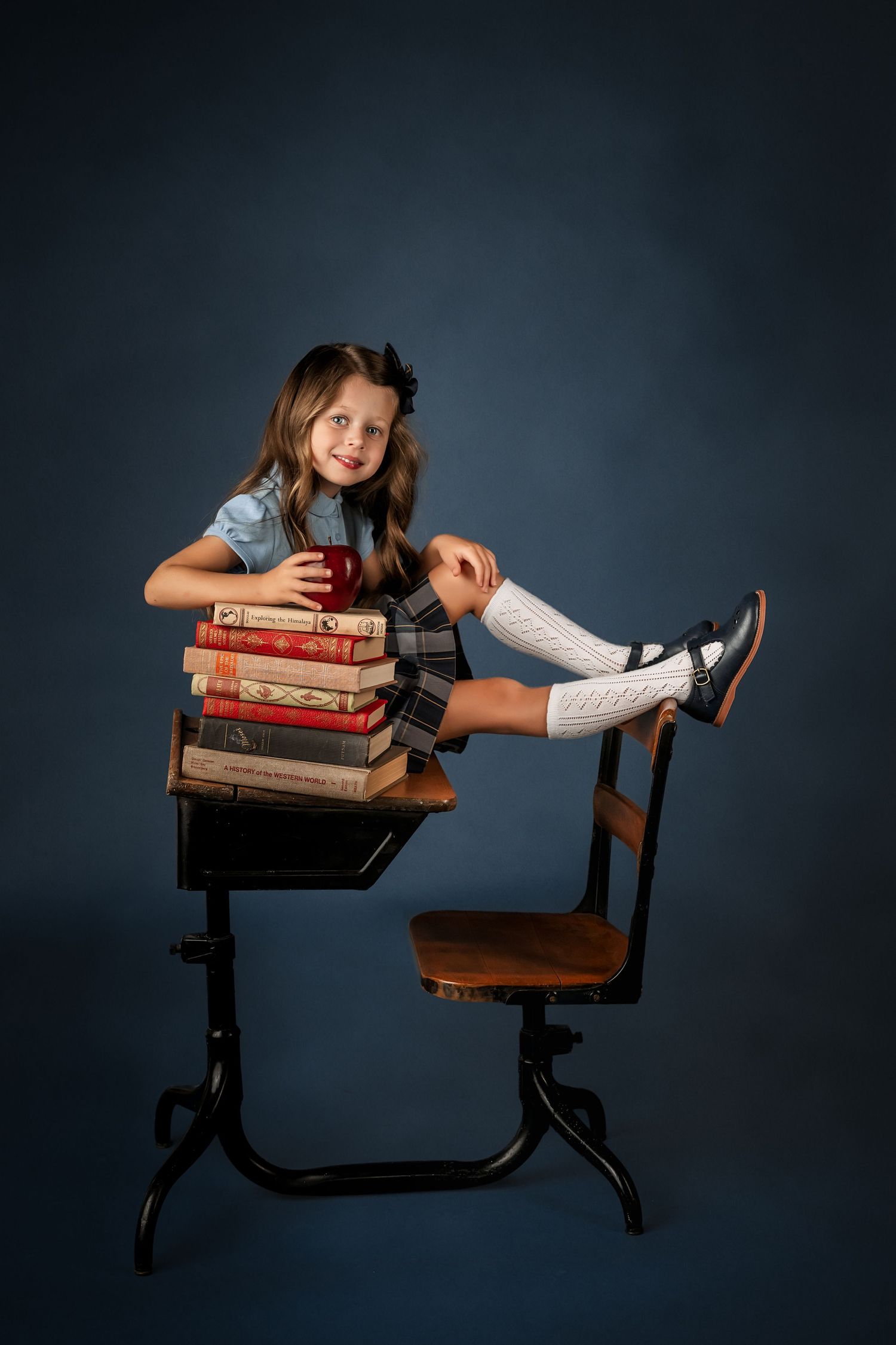Young girl in school uniform sits at vintage desk with stacked books and red apple, posing cheerfully for fine art back to school photos