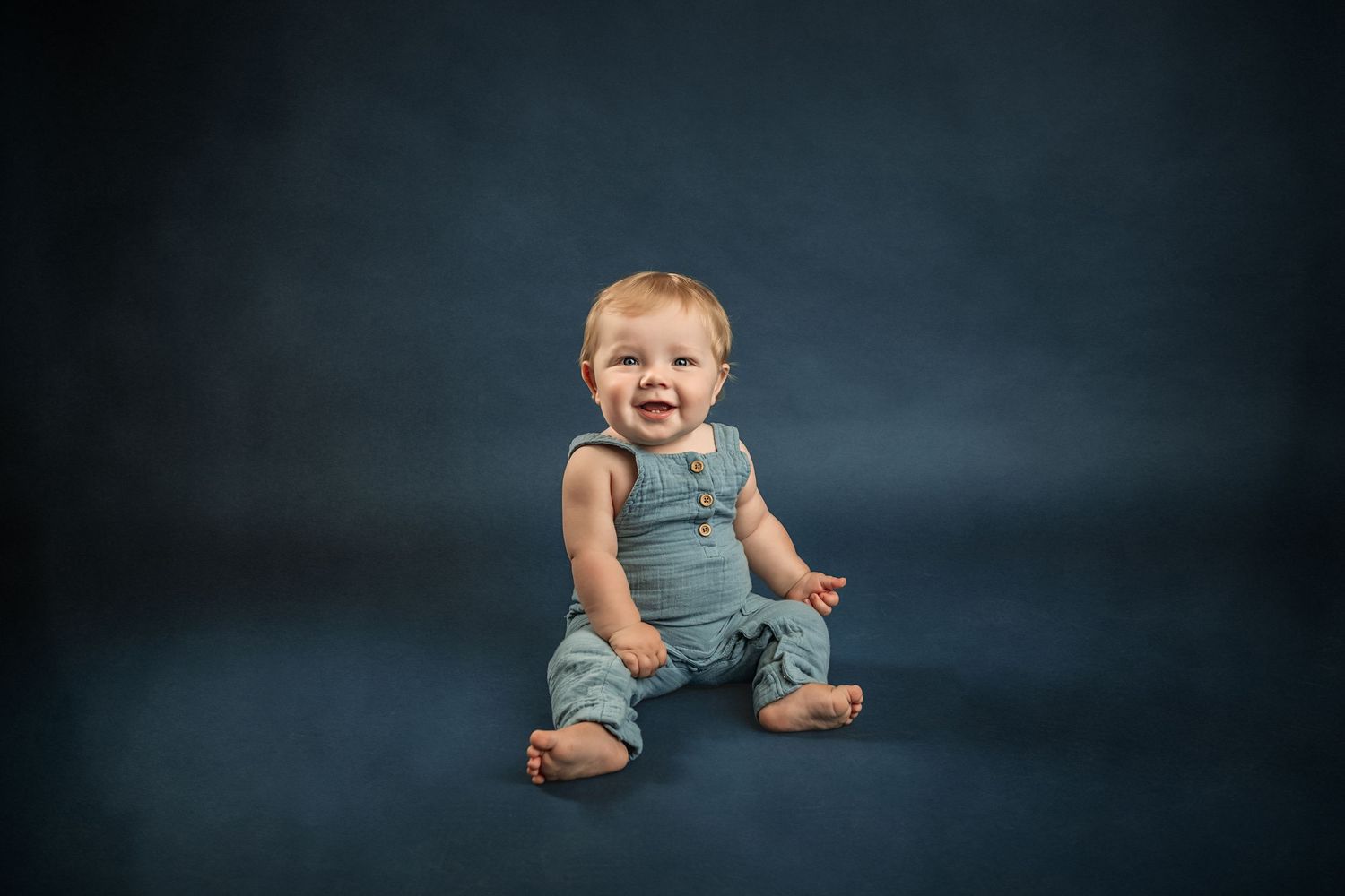 A happy baby in a blue denim romper sits smiling on a dark blue studio background for Marietta fine art children's photographer.