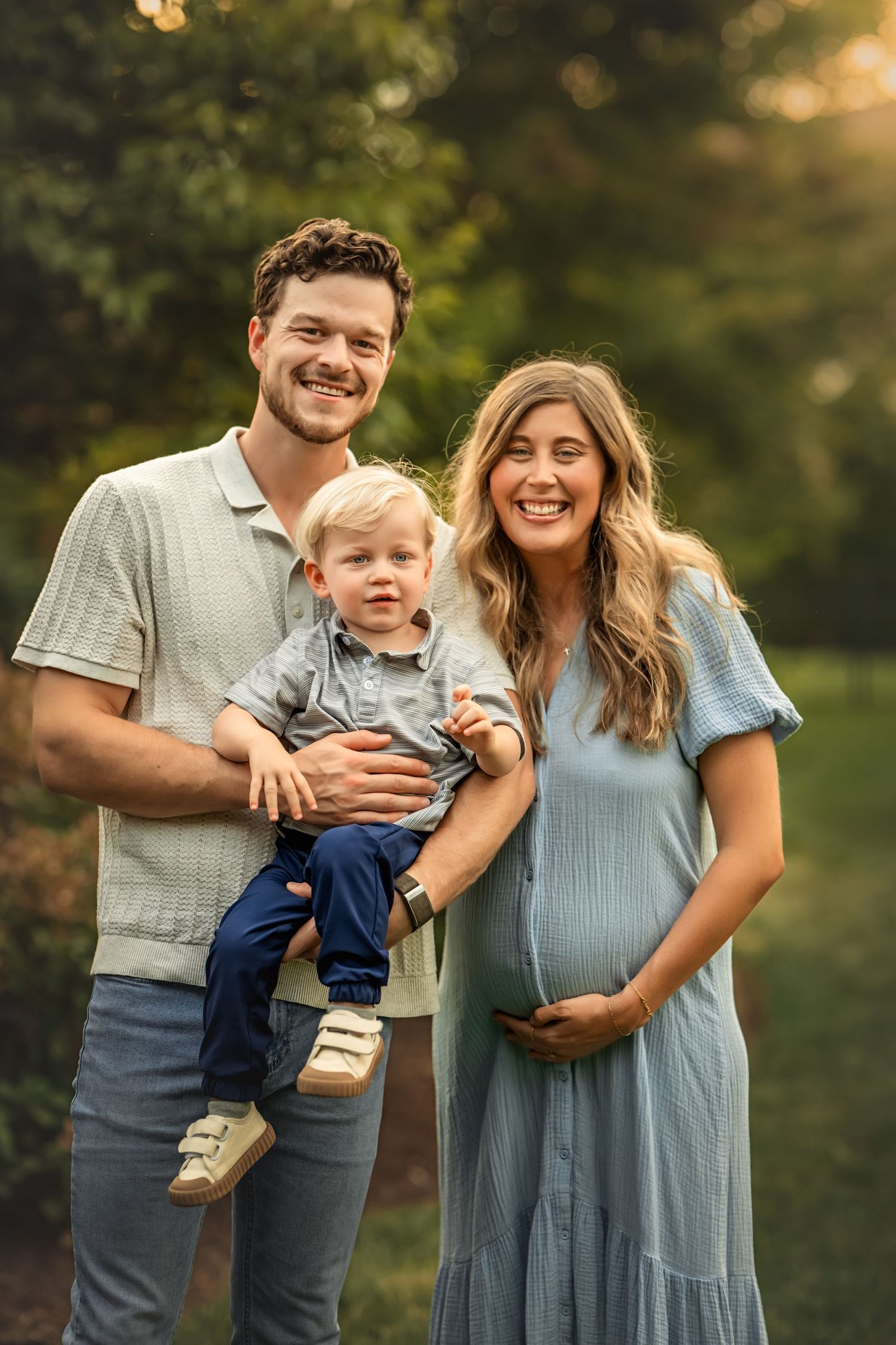 A smiling couple poses outdoors, man holding toddler boy, pregnant woman in blue dress cradling bump, golden hour light.