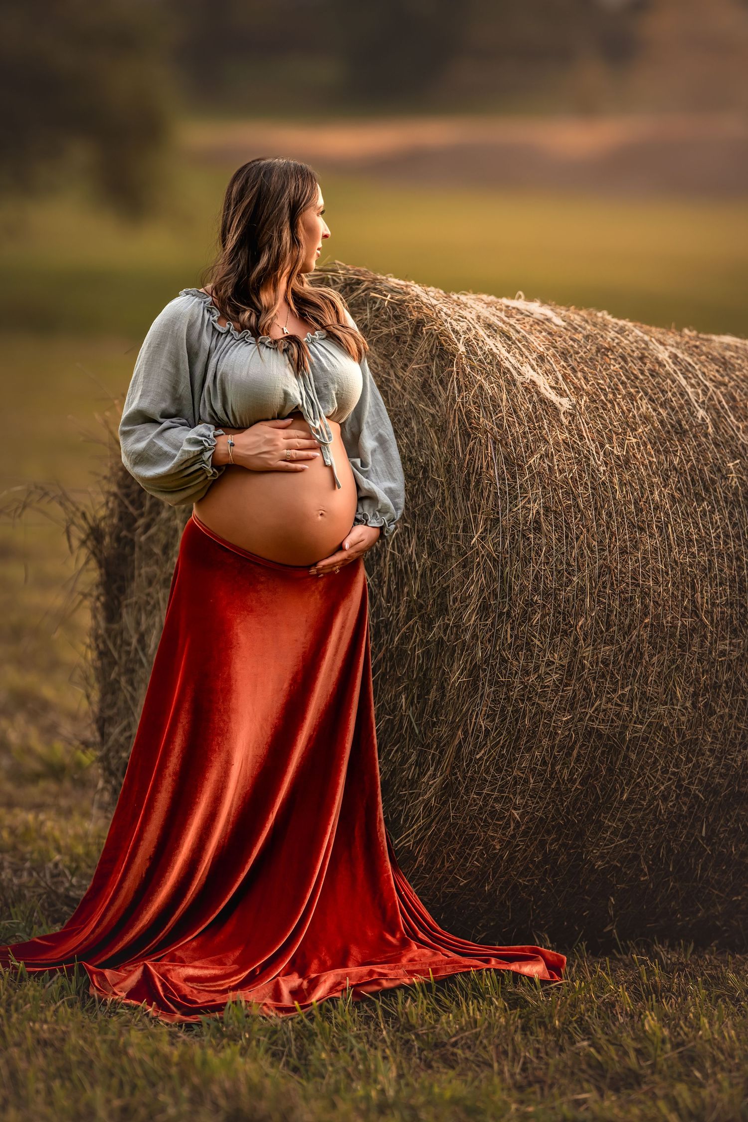 Pregnant woman in red velvet skirt and sage blouse cradling belly beside hay bale in golden field.