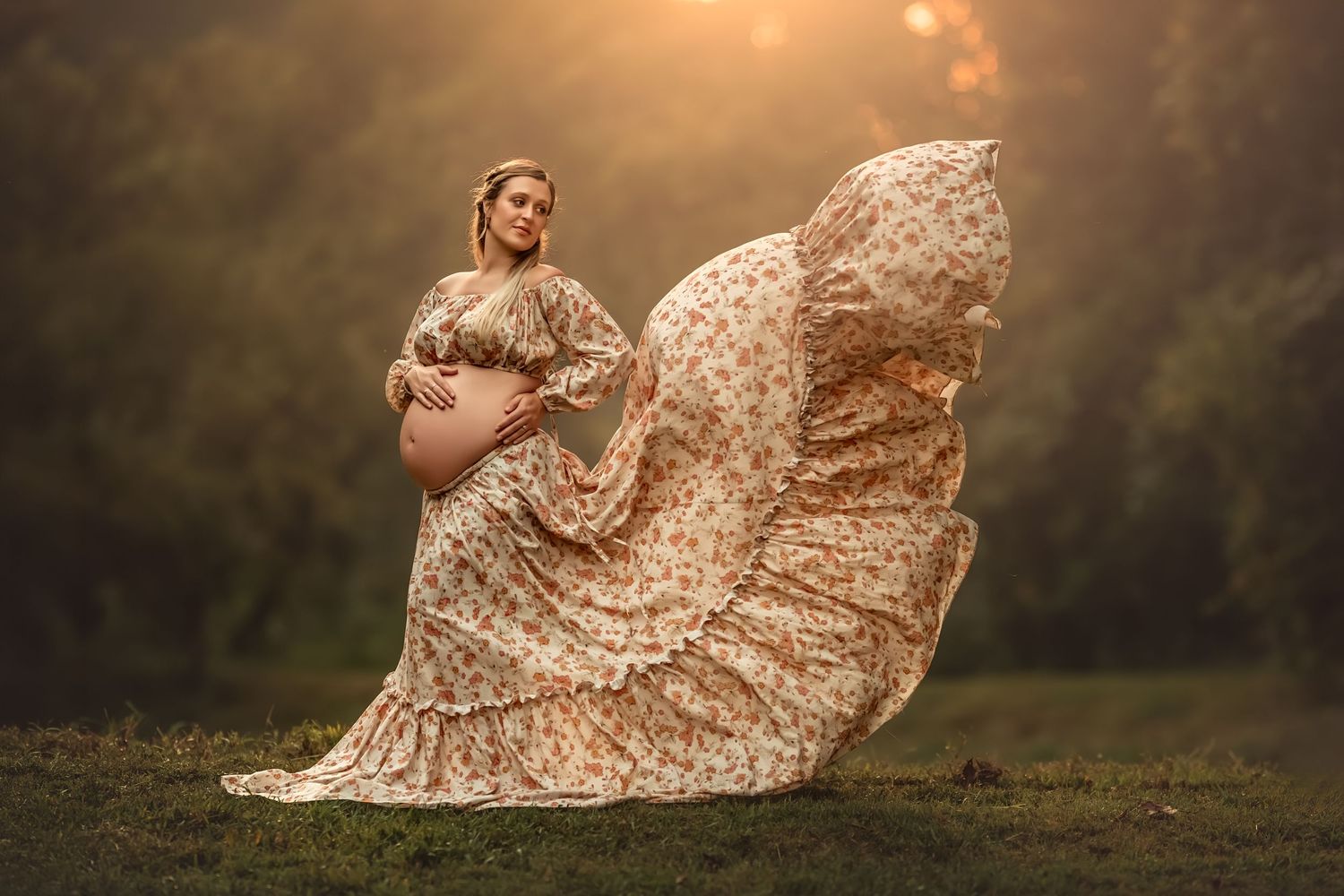 Pregnant woman in flowing floral dress posing outdoors in golden sunset light with fabric billowing in the wind.
