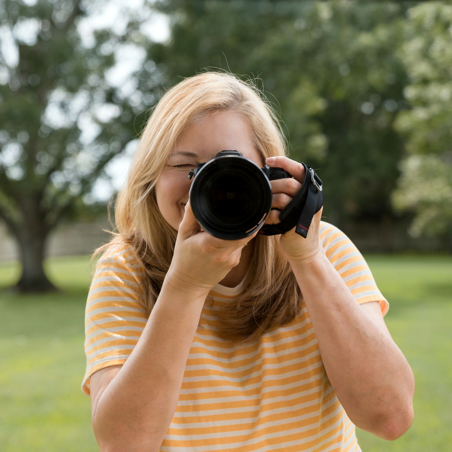 Photographer Kelly J. Russo wearing yellow striped shirt holding professional DSLR camera outdoors in sunny garden setting.