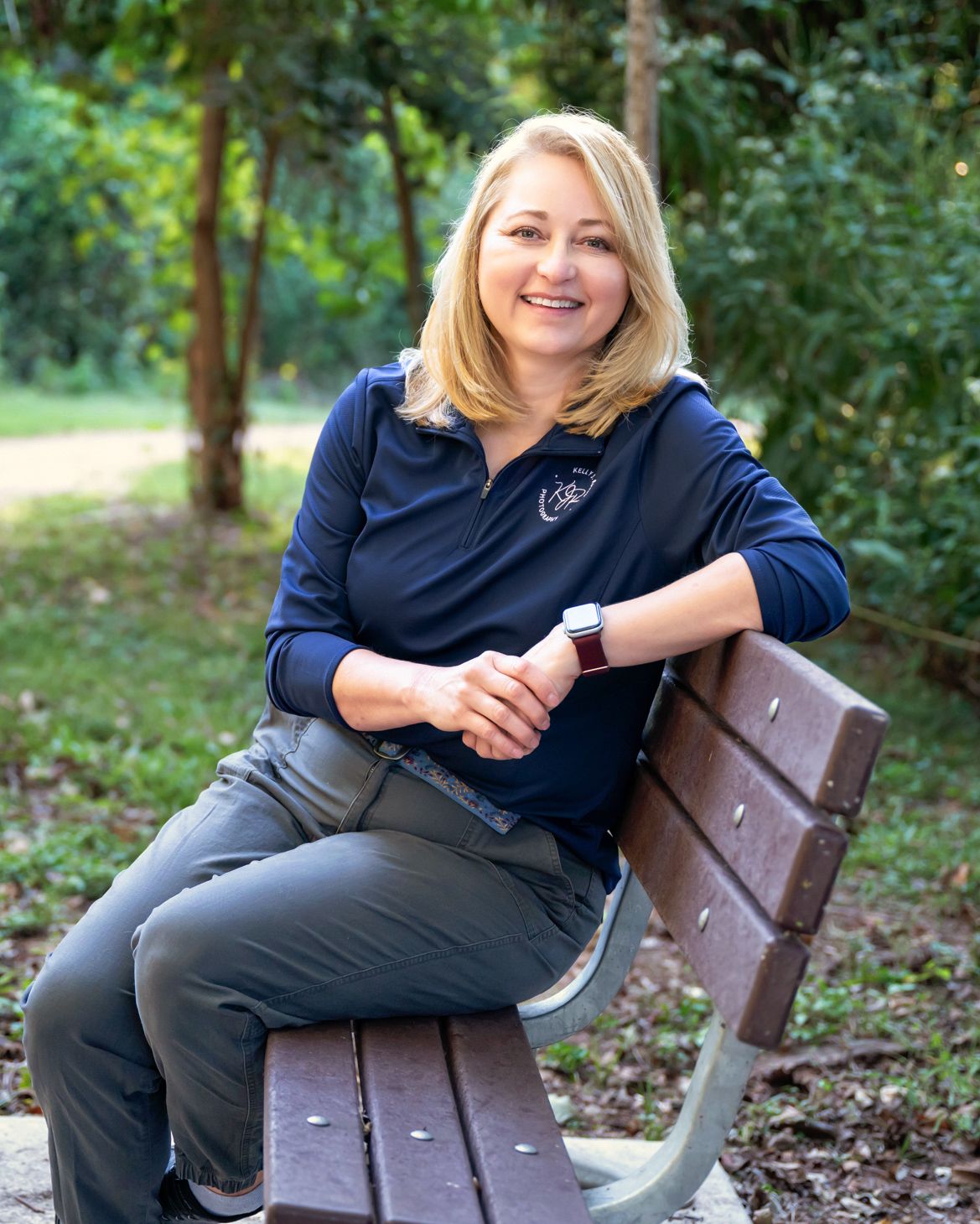 Photographer Kelly J. Russo wearing a navy blue polo shirt sits smiling on a wooden park bench surrounded by green trees.