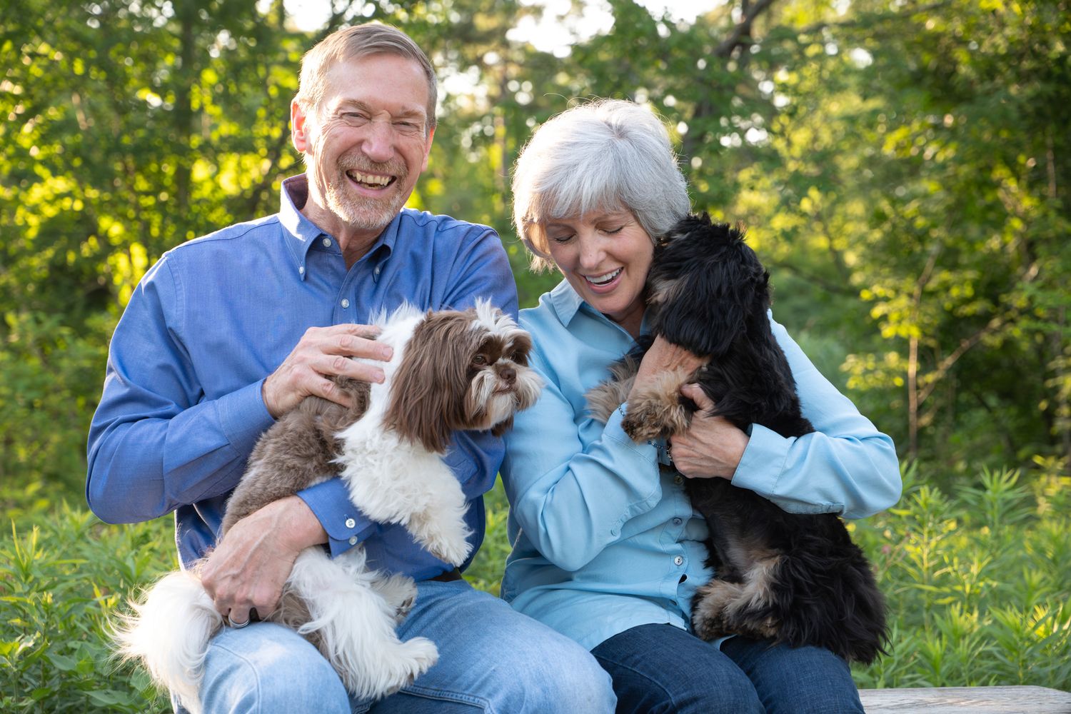 A happy couple sits outdoors in a garden setting with their two small dogs, enjoying a sunny afternoon.