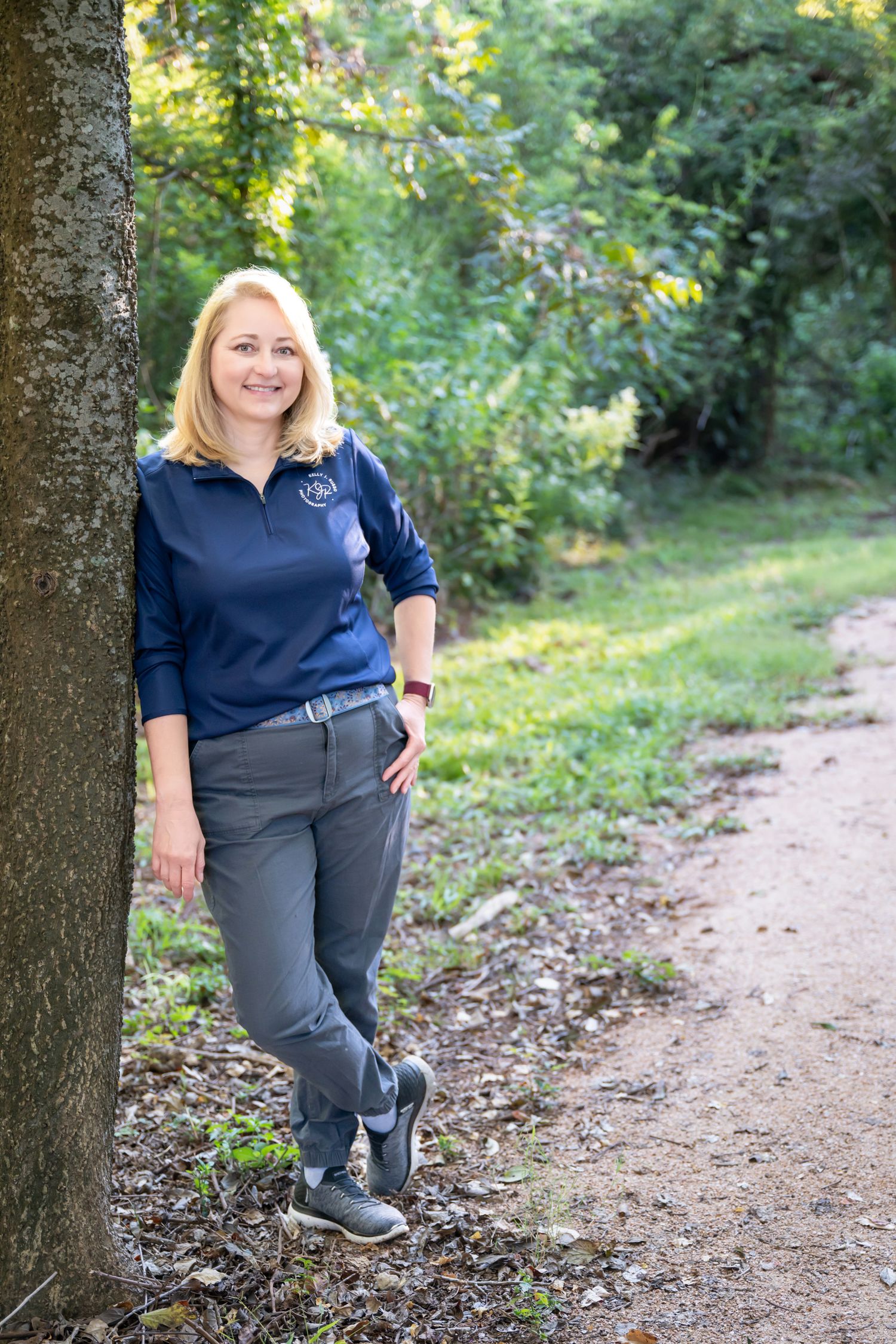 Kelly J. Russo stands casually leaning against a tree trunk along a dirt path in a green wooded area on a sunny day.