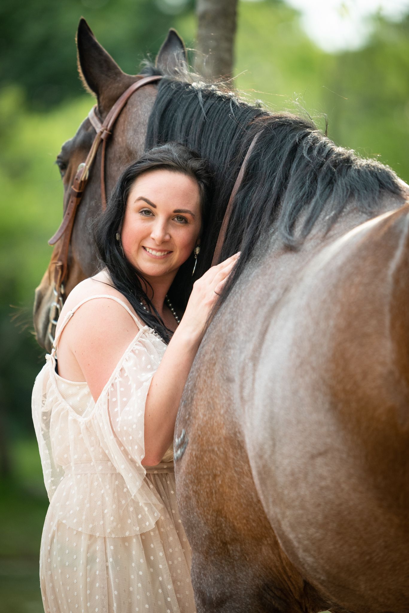 A person in a lace dress poses affectionately with a large brown horse against a blurred green background.