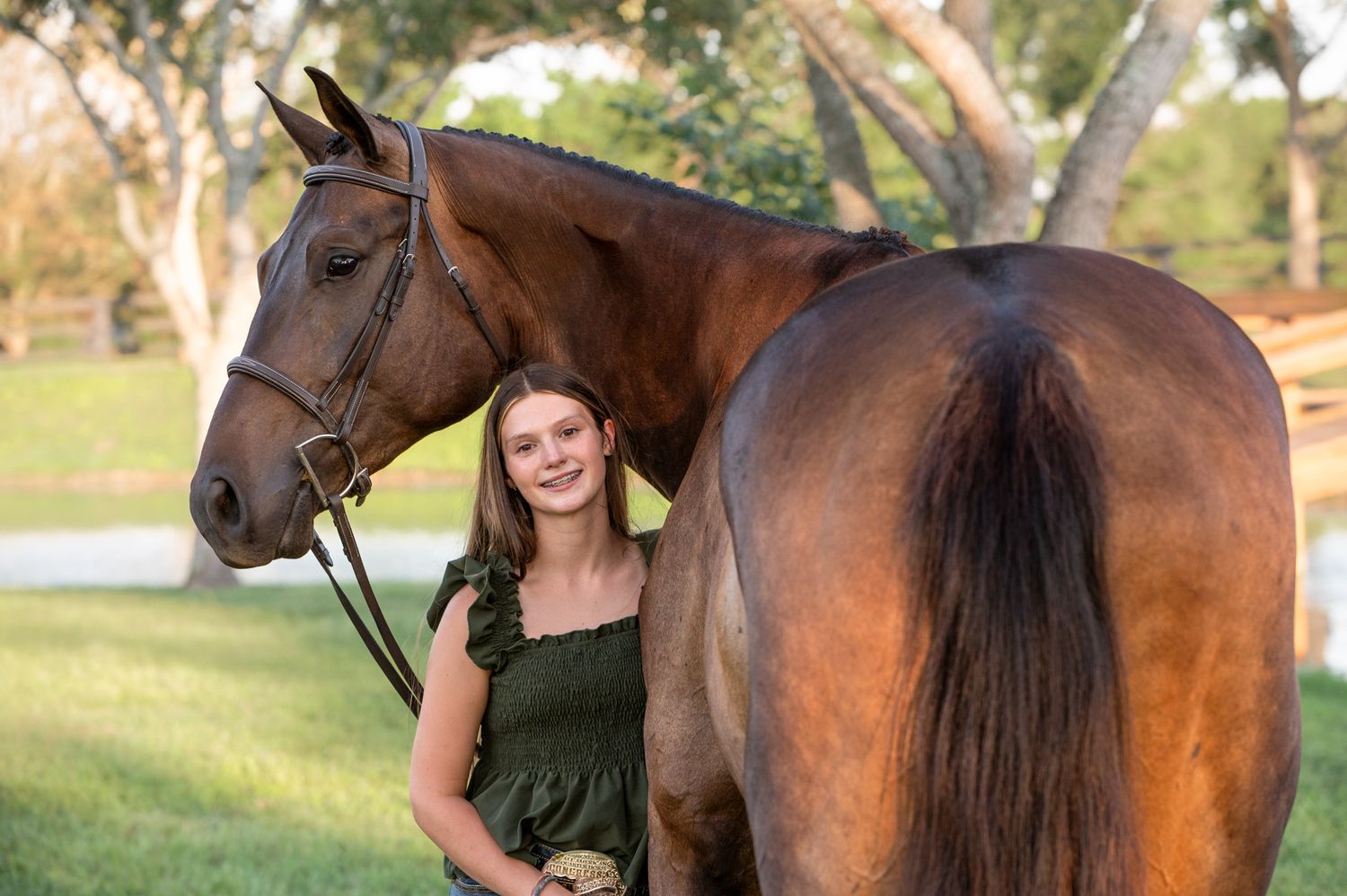 A bay horse stands in profile on grass while someone in a green top poses beside it in warm sunlight.