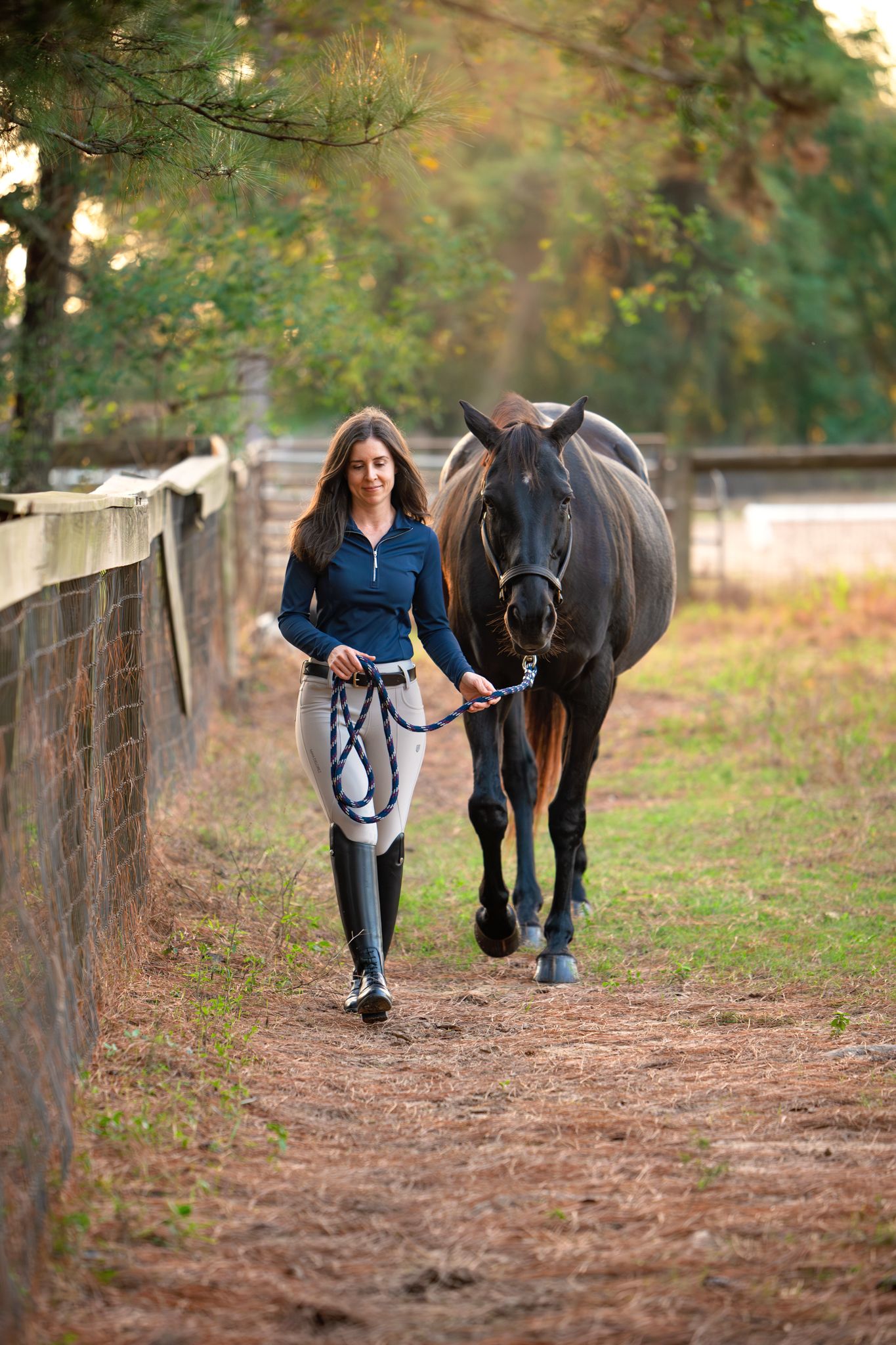 A dark horse and person in blue walking together along a tree-lined dirt path on an autumn afternoon.