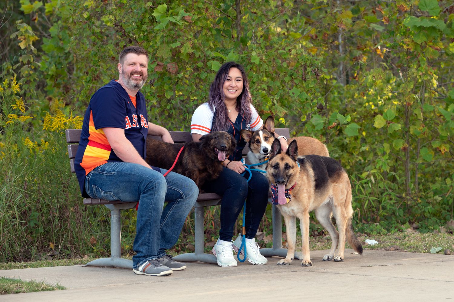 Two people sit on a bench with a large German Shepherd dogs and two corgis on a nature trail surrounded by greenery.