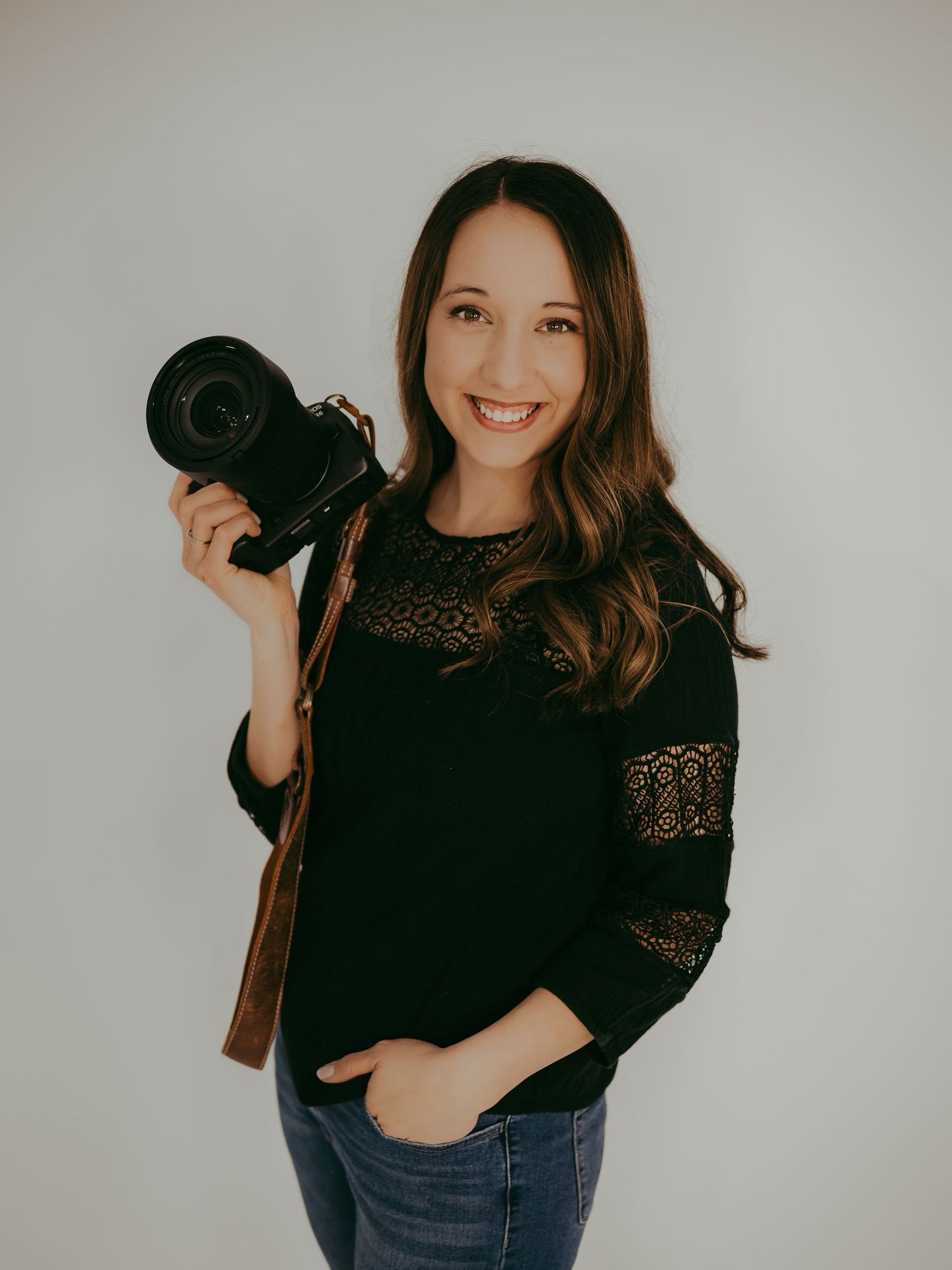 A photographer in a black lace sweater and jeans holds a professional camera against a light gray studio background.
