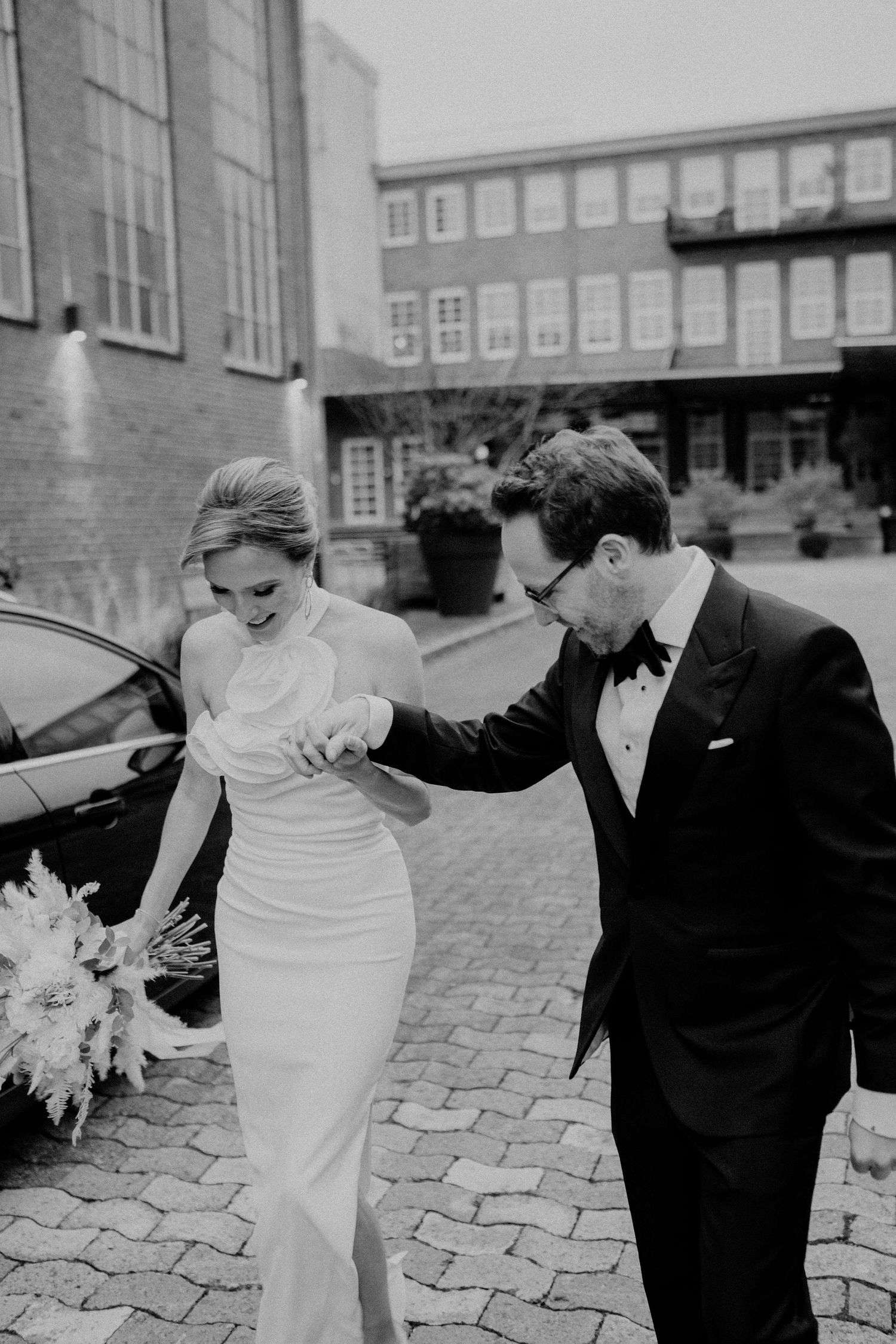 A bride and groom in classic black and white attire walk together on cobblestone street holding hands.