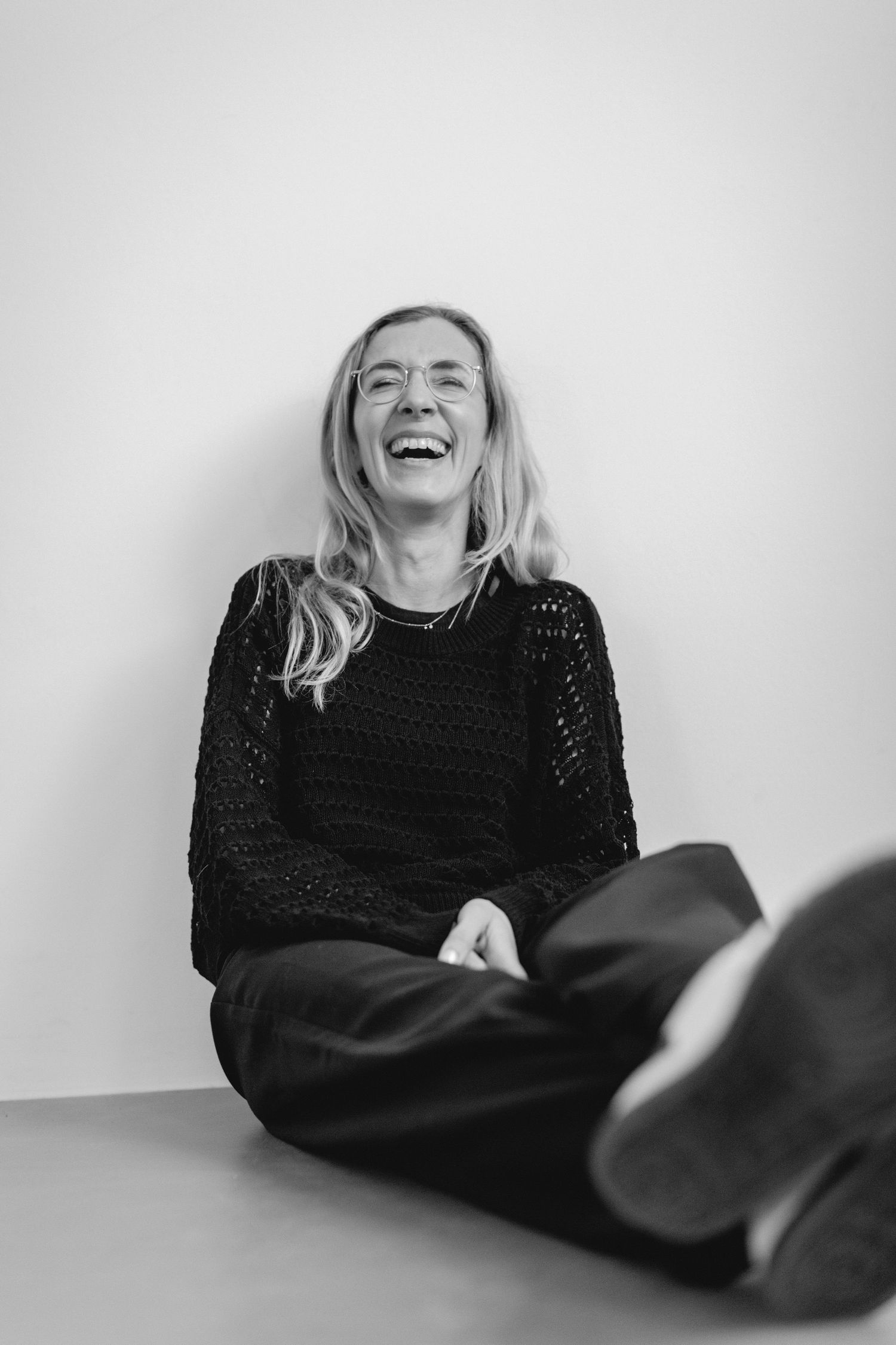 A black and white portrait of a photographer captures a joyful moment as someone wearing a dark sweater laughs while sitting against a wall.