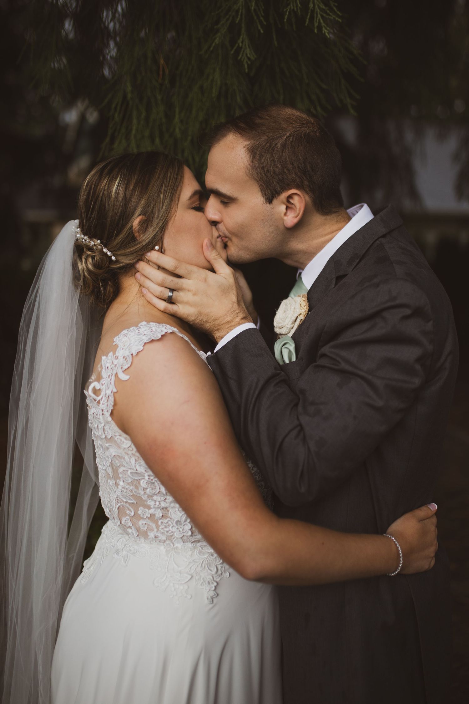 A romantic outdoor wedding moment captured between a bride in a lace gown with veil and groom in dark suit.