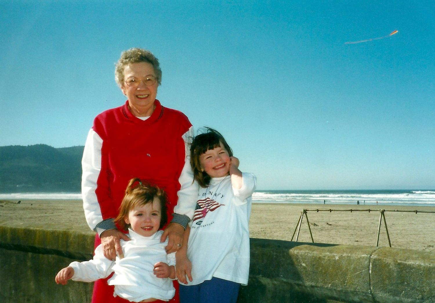 Three people pose together at the beach on a sunny day with mountains and blue sky in the background.