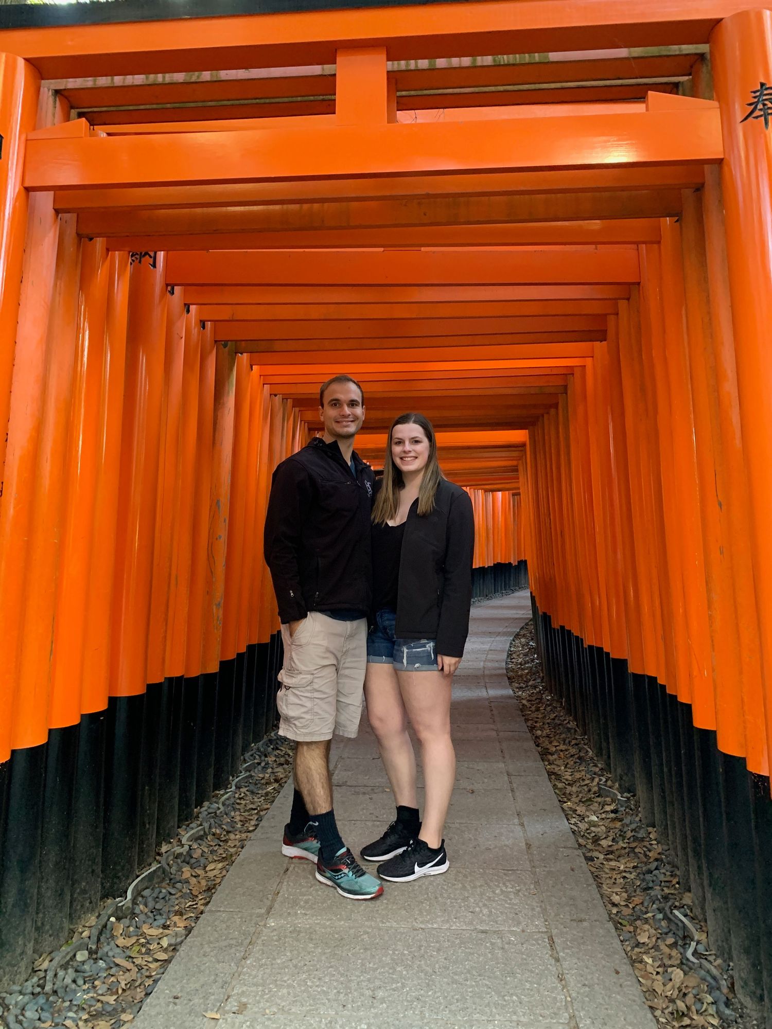 Two tourists pose in the iconic orange torii gates path at Fushimi Inari Shrine in Kyoto, Japan.