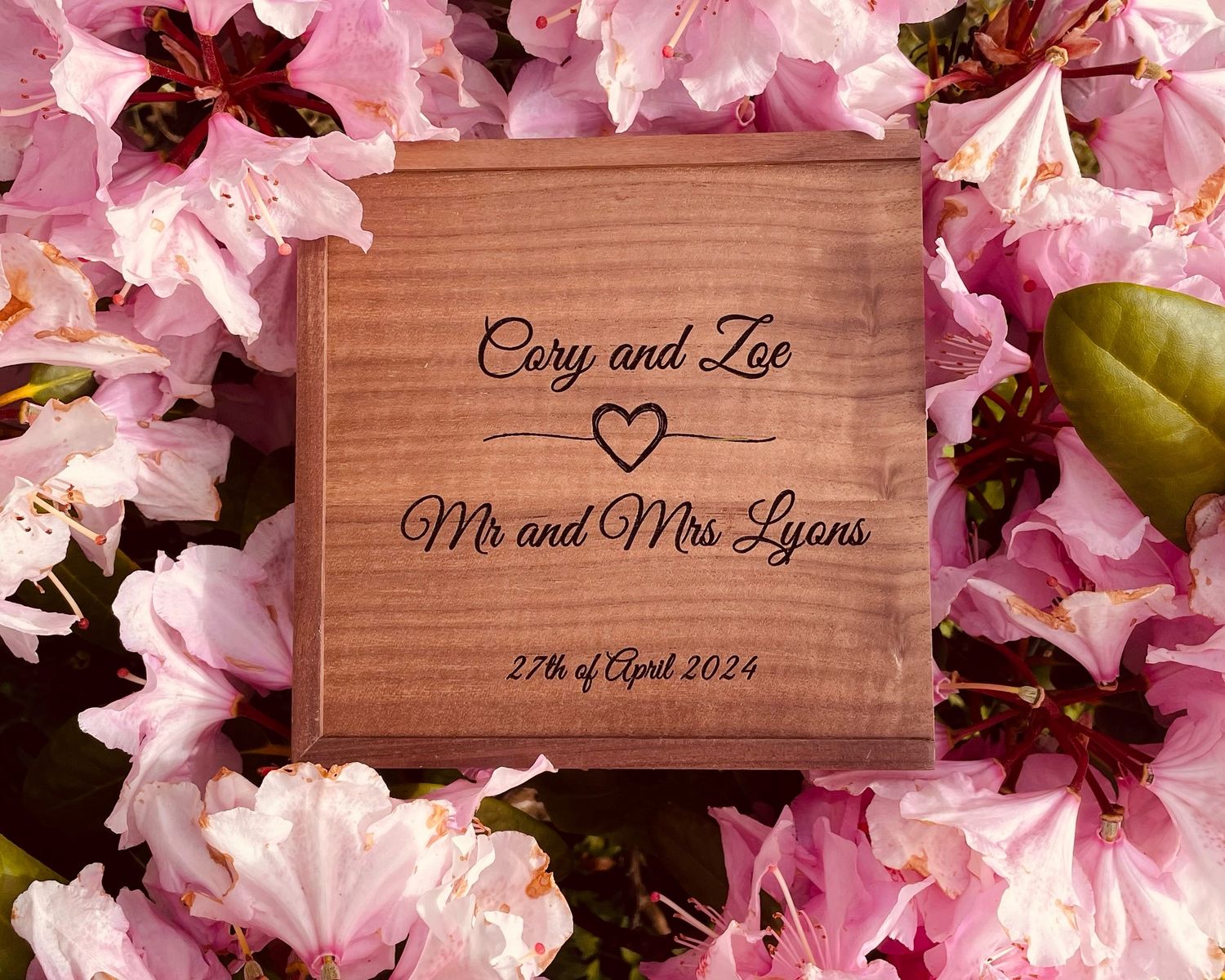 Custom engraved wooden box nestled among pink azalea flowers displaying wedding details.