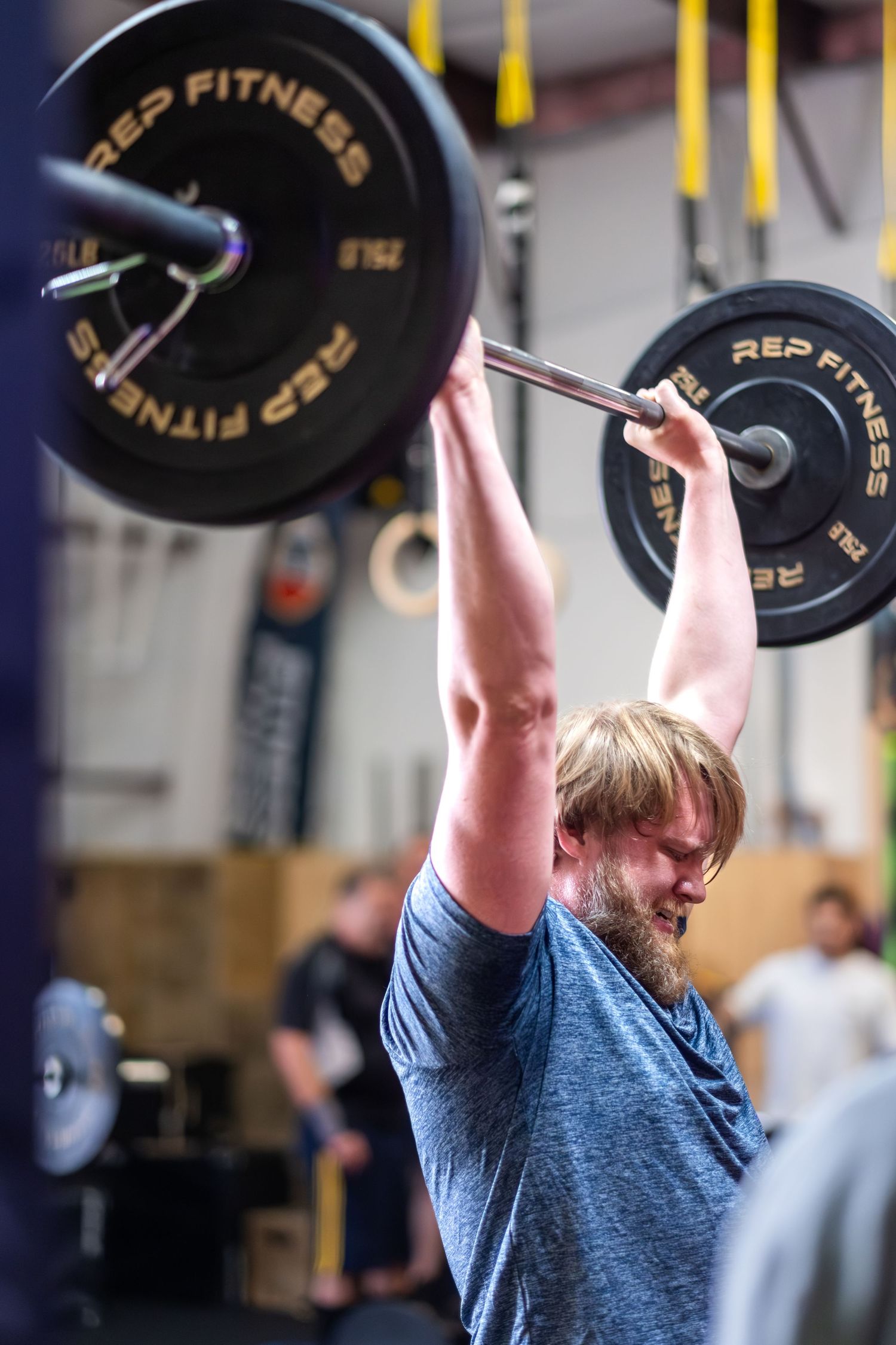 Bearded man pressing heavy REP Fitness barbell overhead during CrossFit workout.