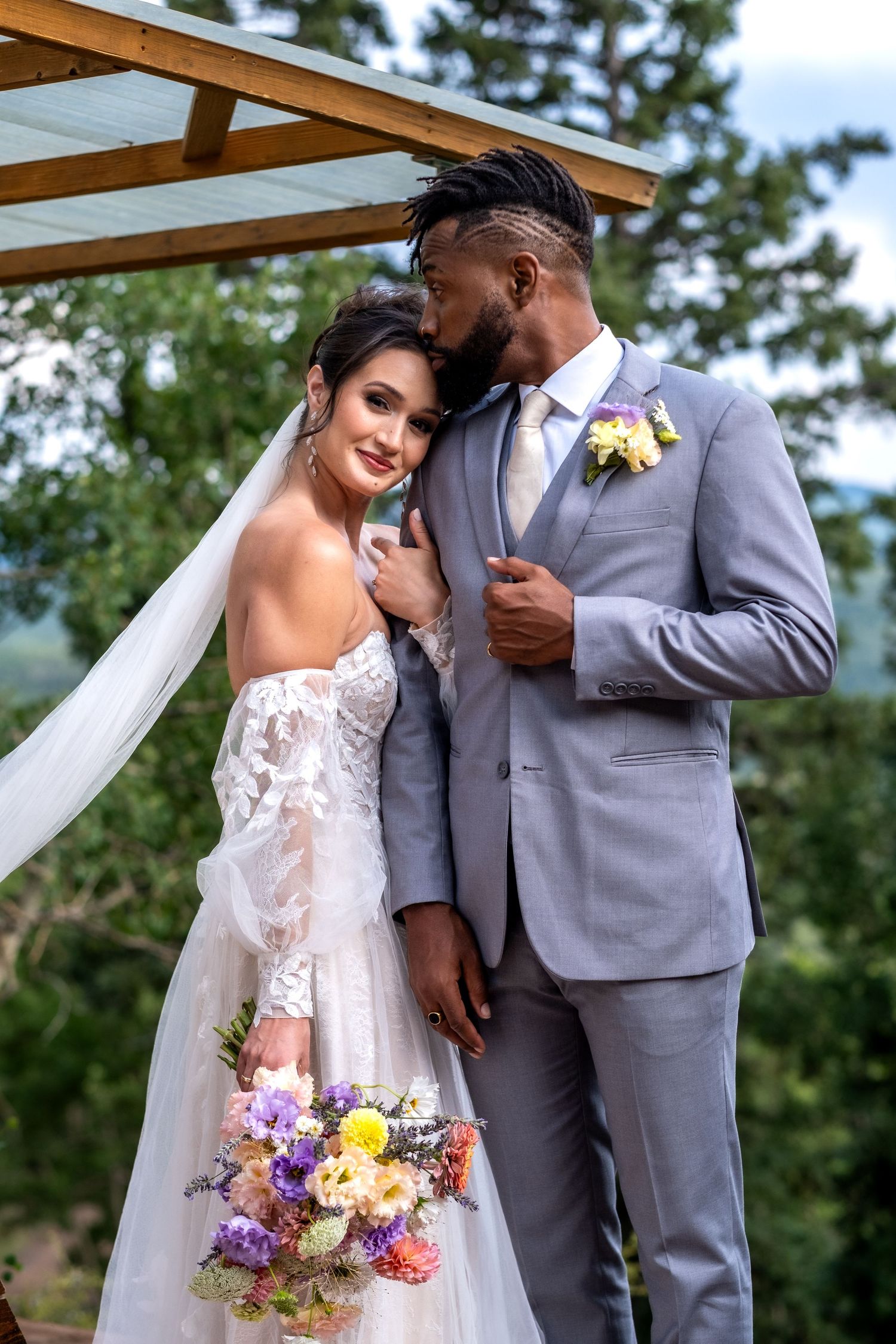 Bride and groom posing under a pergola, bride holding colorful bouquet in a lace off-shoulder gown.
