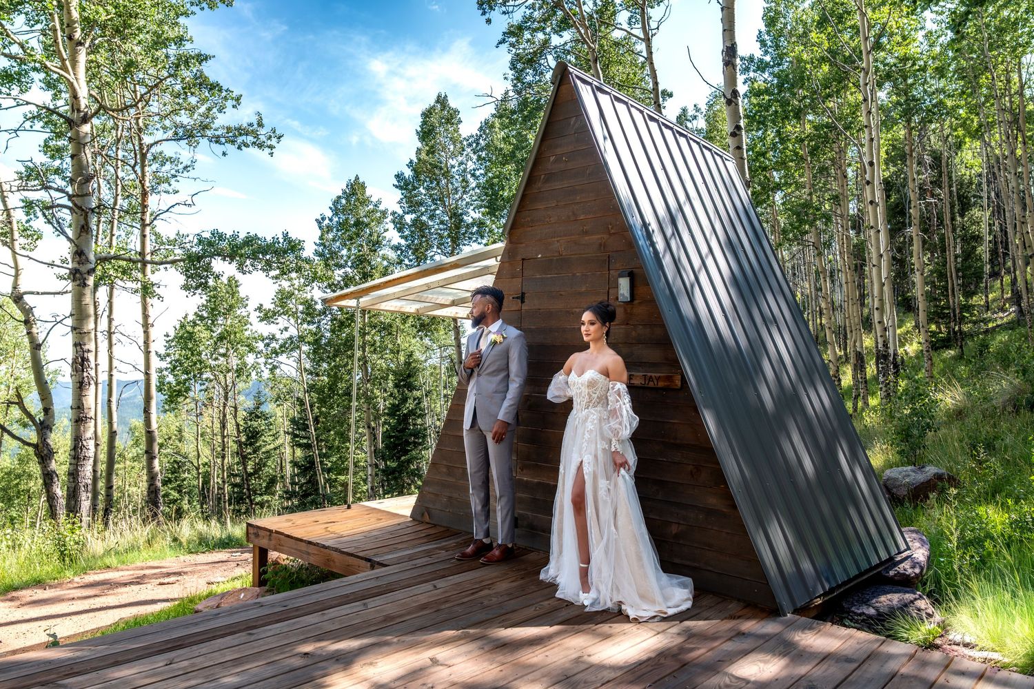Bride and groom standing on wooden deck of A-frame cabin surrounded by lush forest trees.