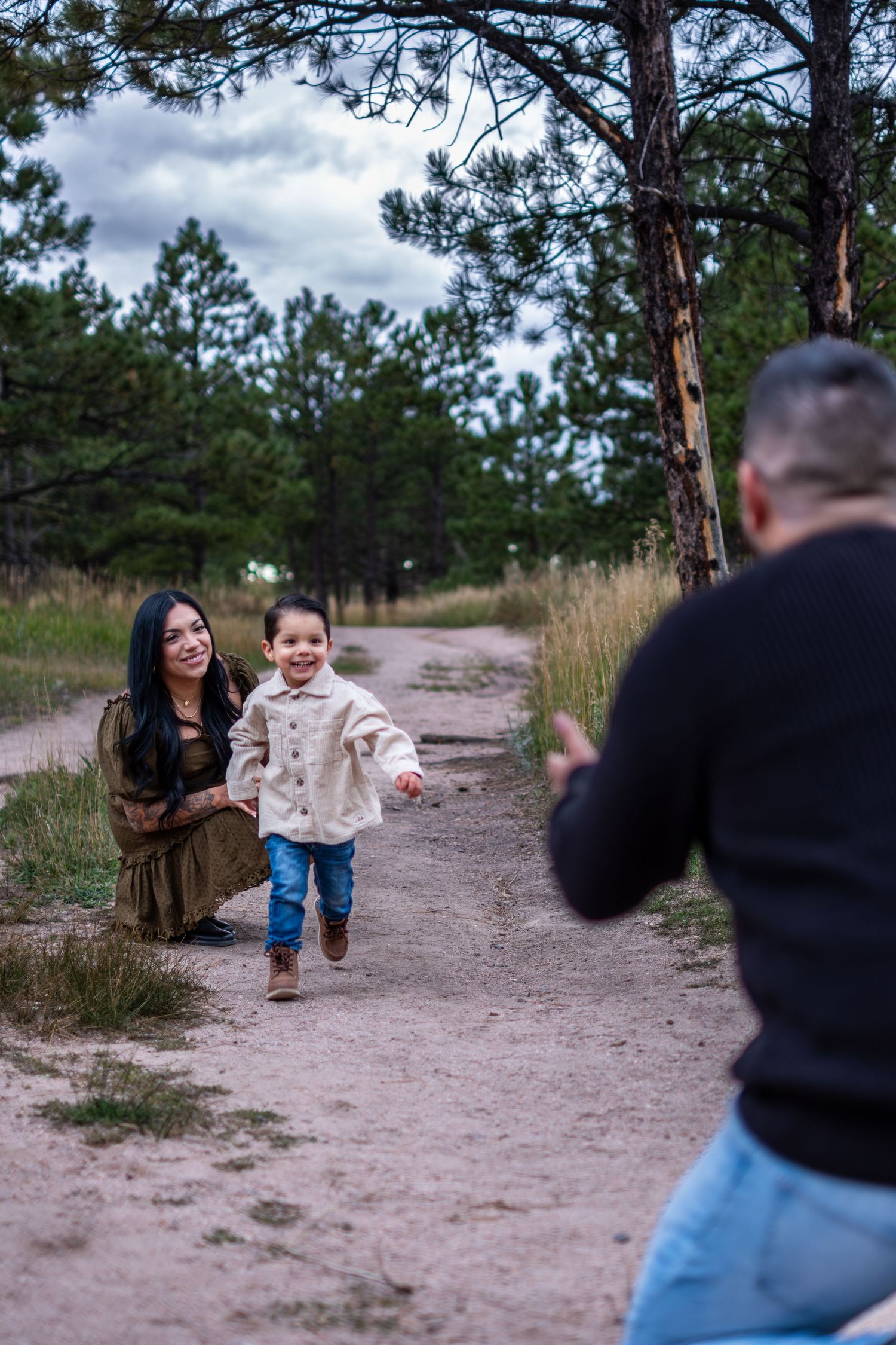 Smiling toddler running toward father on dirt path as mother watches in pine forest.