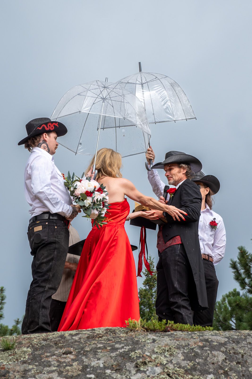 Western wedding ceremony on rocky hilltop with bride in red gown holding bouquet under clear umbrella in overcast sky.