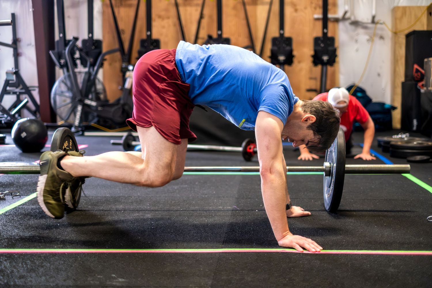 Male athlete performing a burpee over a barbell on a gym floor during a CrossFit workout.