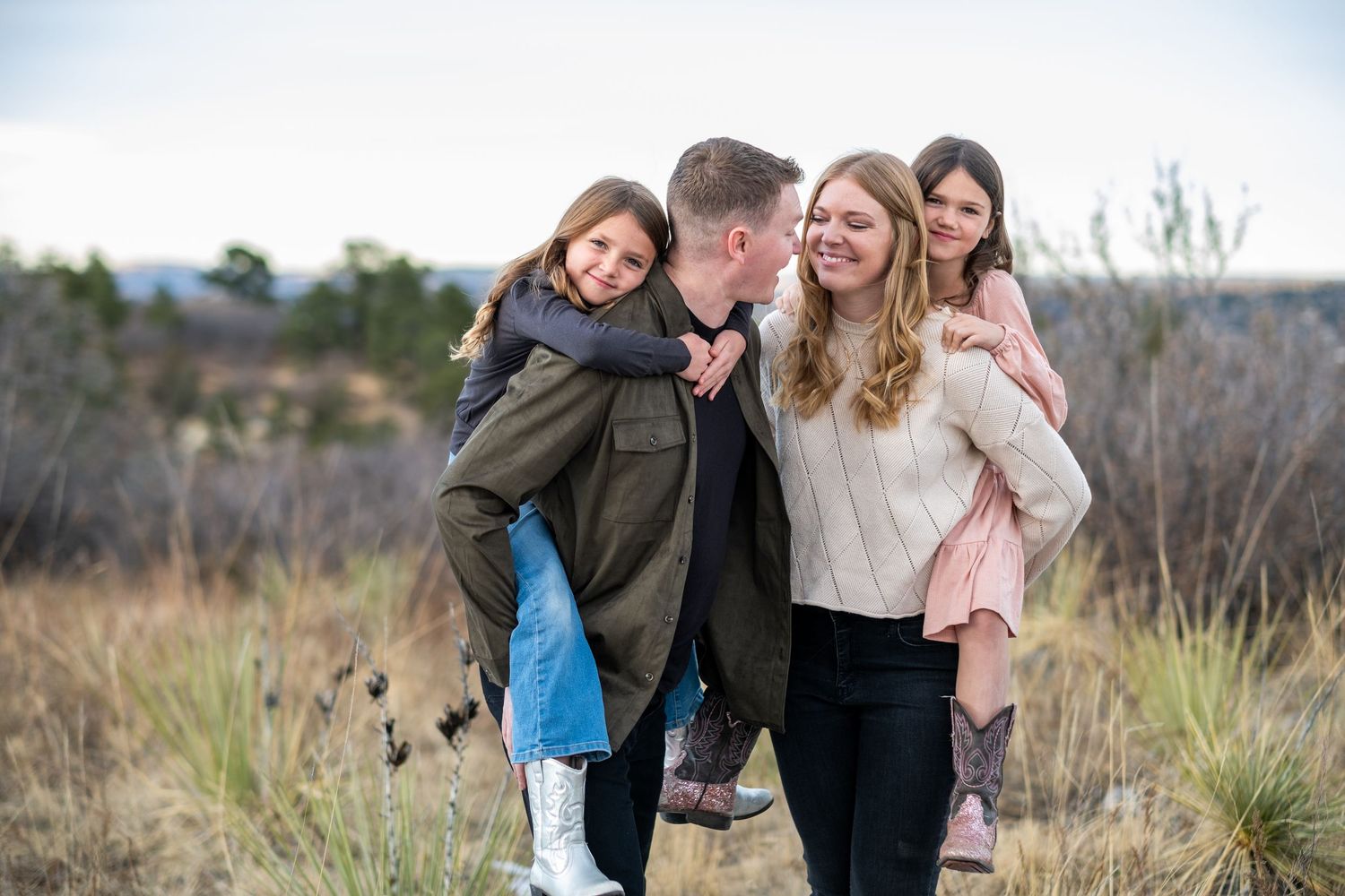 Joyful family walking outdoors, father and mother giving piggyback rides to two laughing daughters in field.