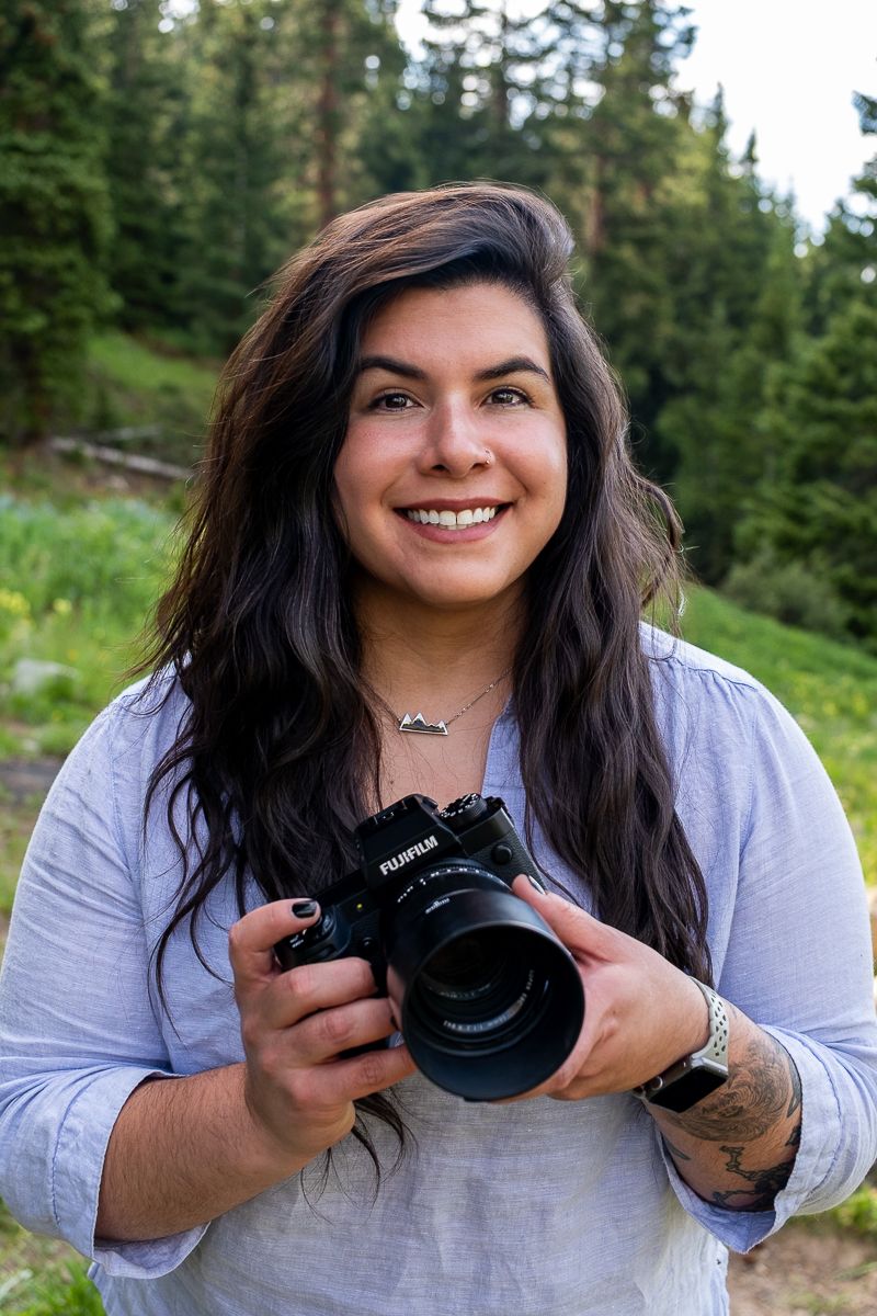 Smiling female photographer holding a Fujifilm camera outdoors in a green forest setting.