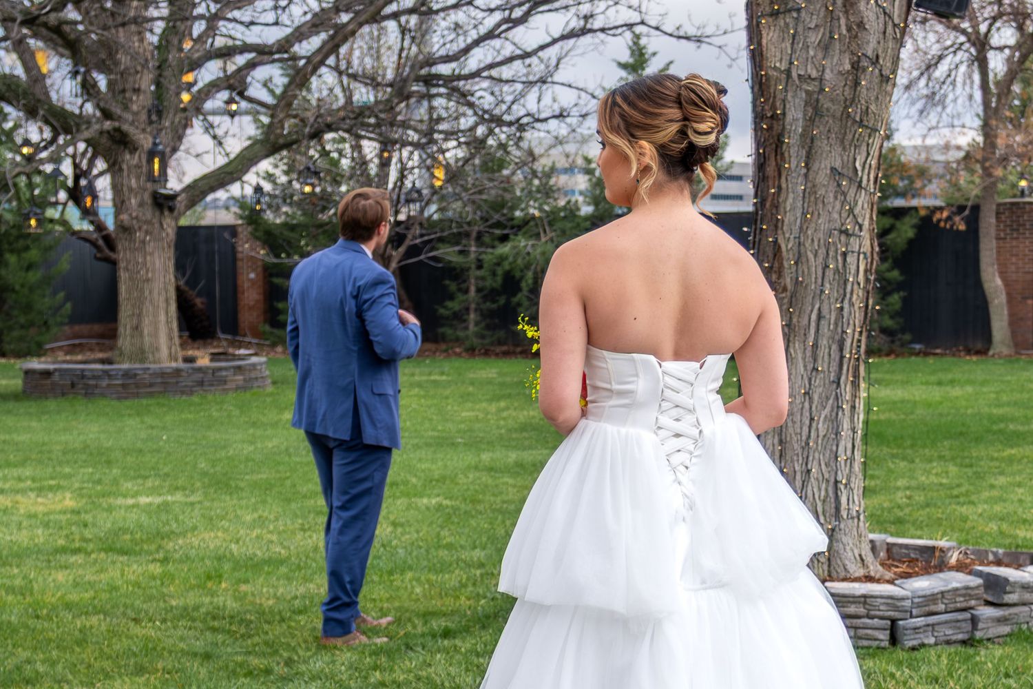 Bride in strapless corset gown awaiting first look with groom in blue suit outdoors.