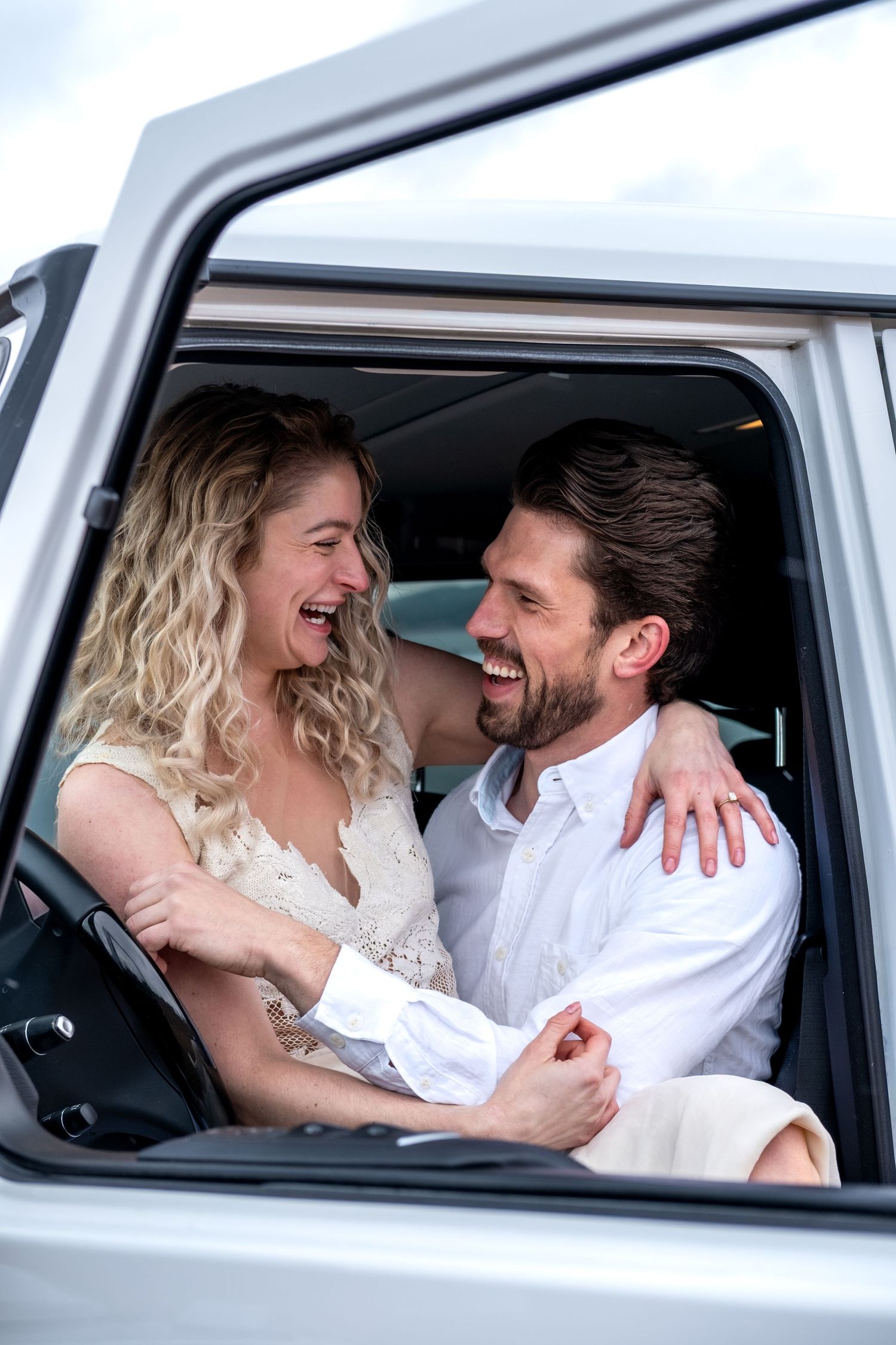 Happy couple laughing and embracing inside a white vehicle, dressed in wedding attire on their special day.