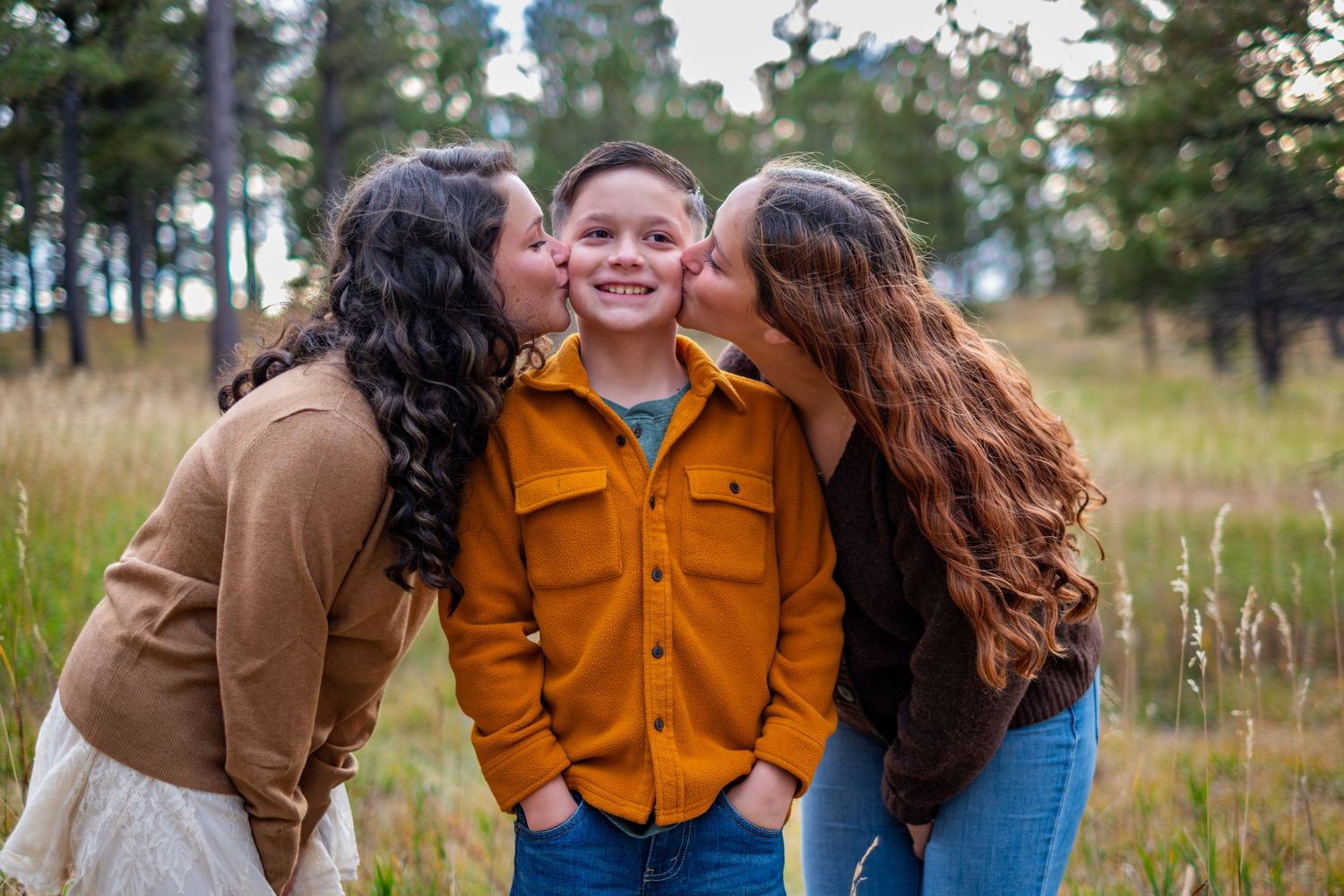 Two sisters kissing their smiling brother on the cheeks outdoors in a forest meadow.