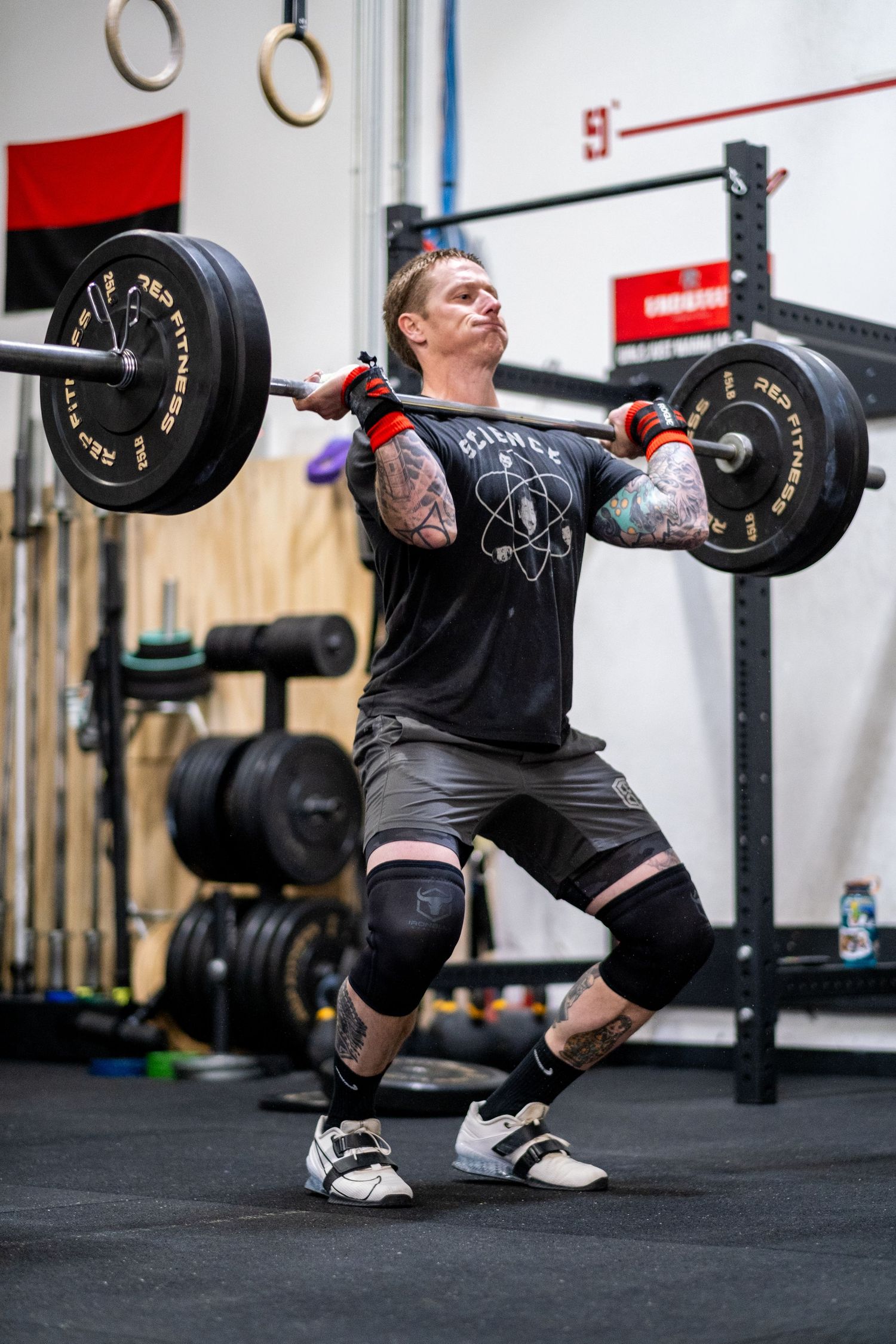 Tattooed man performs a barbell clean exercise wearing knee sleeves in an indoor CrossFit gym.