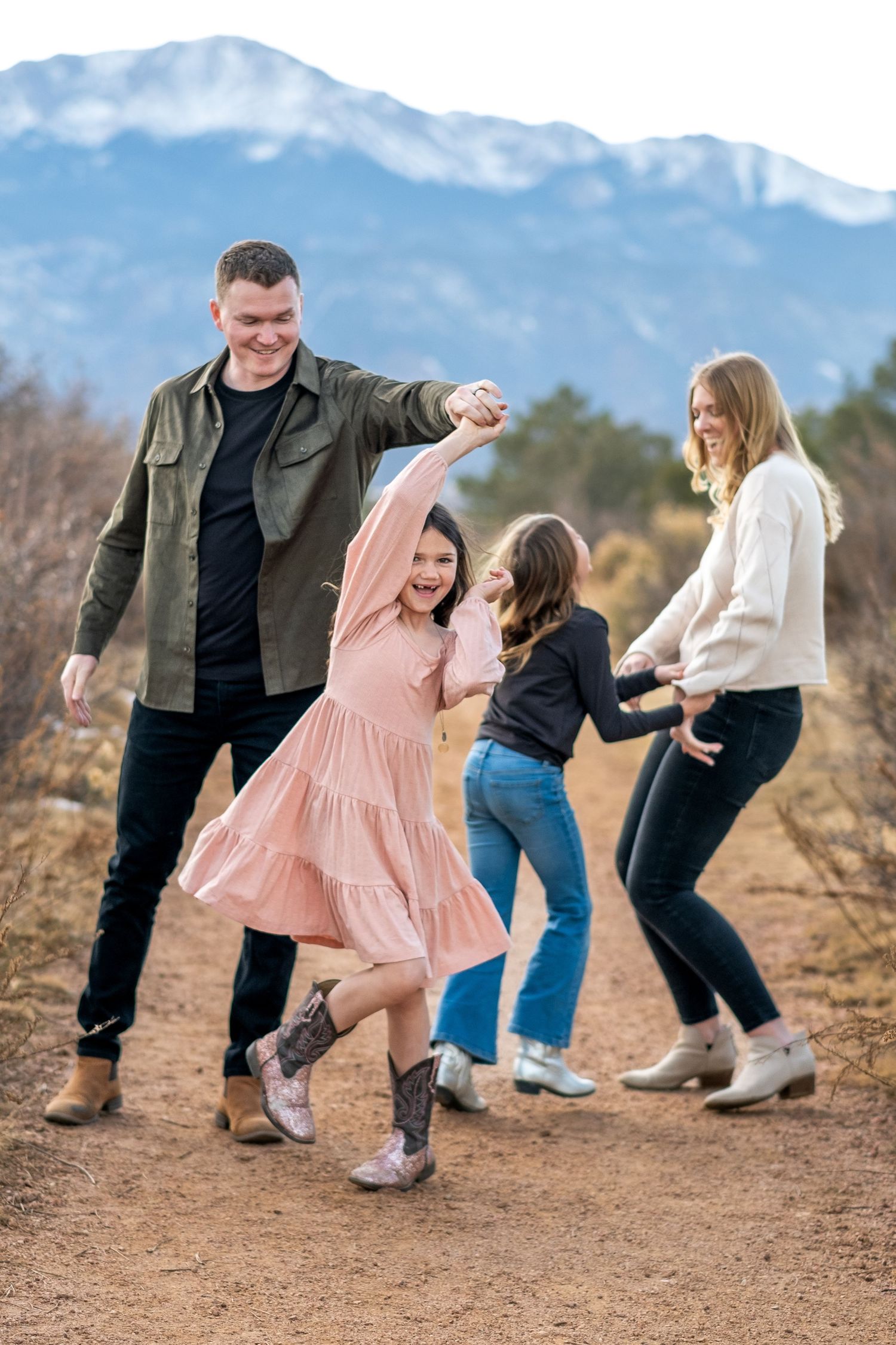 Family of four dancing together on a dirt path with mountain views in a fun outdoor photo session.