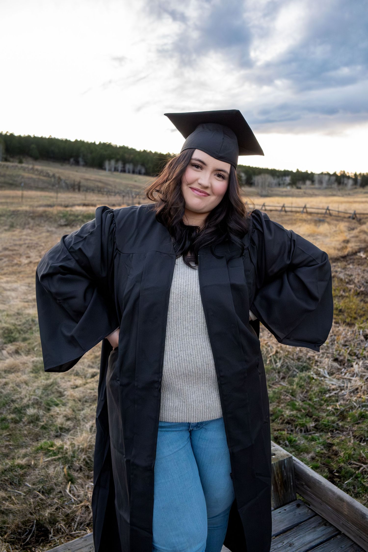 Smiling graduate in black cap and gown with hands on hips posing in scenic outdoor field setting.