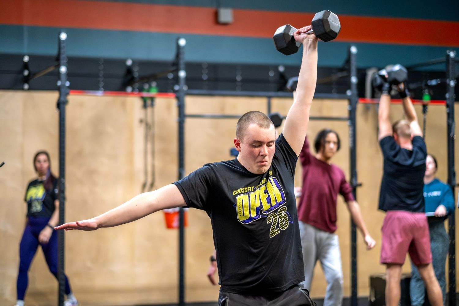 Man in CrossFit Open shirt performs a dumbbell snatch overhead in a busy CrossFit gym.