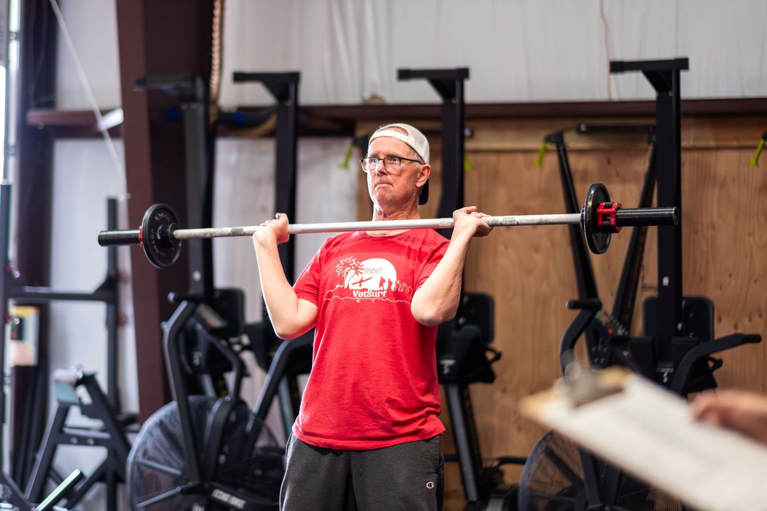 Middle-aged man in red shirt doing barbell squats in a gym setting.