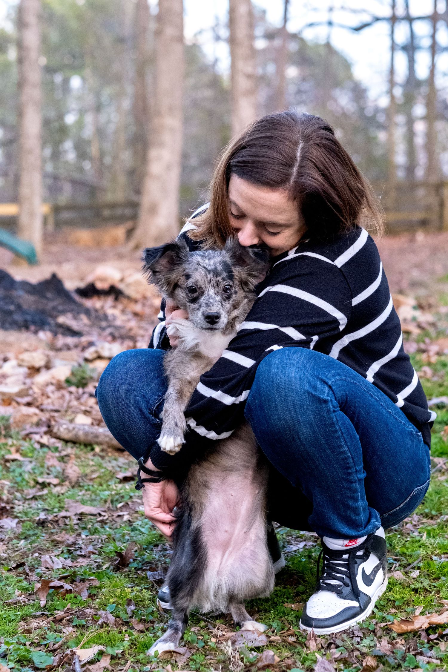 Young woman crouching outdoors hugging a small merle Australian Shepherd puppy in a wooded area.