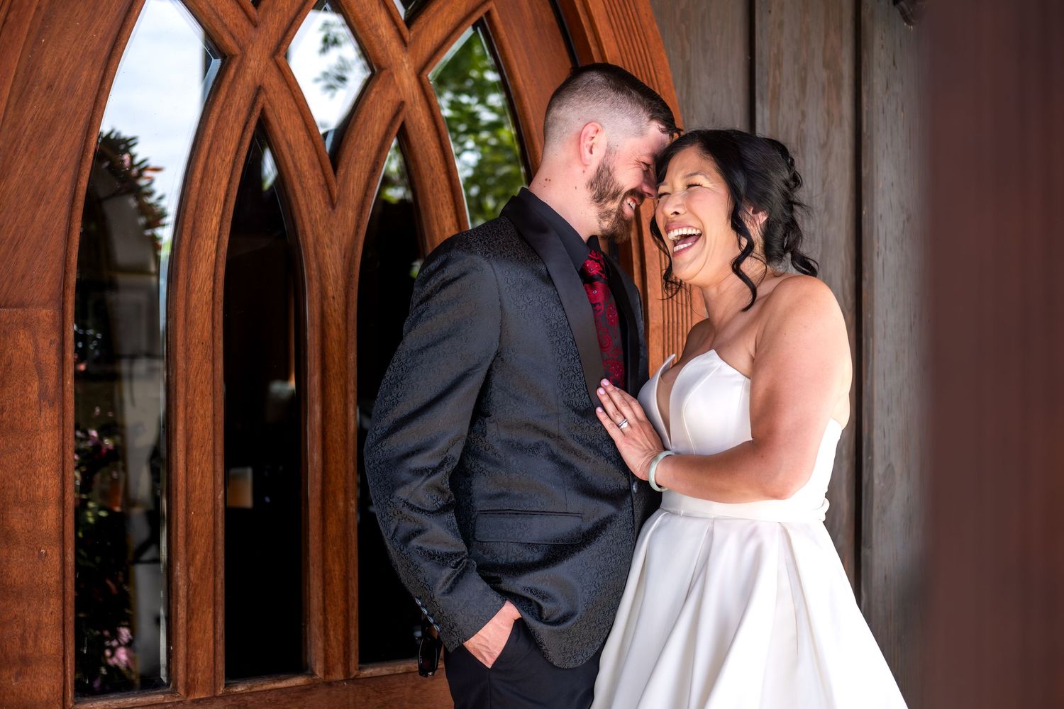 Laughing bride in strapless gown embraces groom in dark suit before elegant wooden church doors.