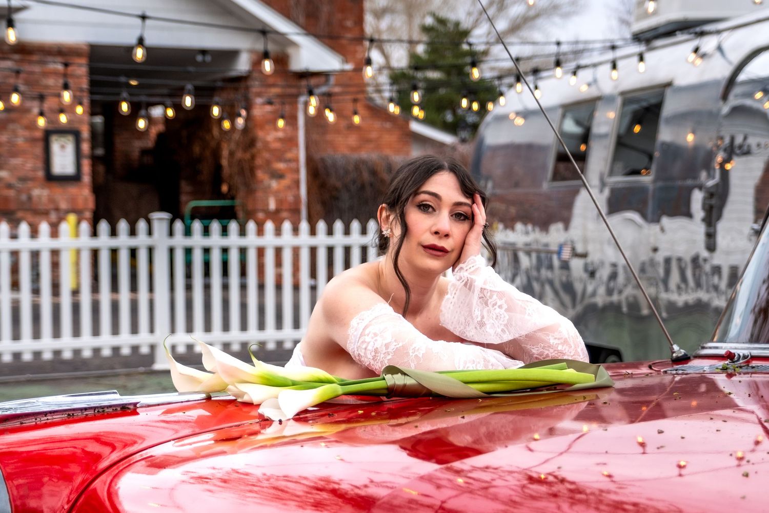 Elegant bride in lace gown leaning on vintage red car hood under string lights.