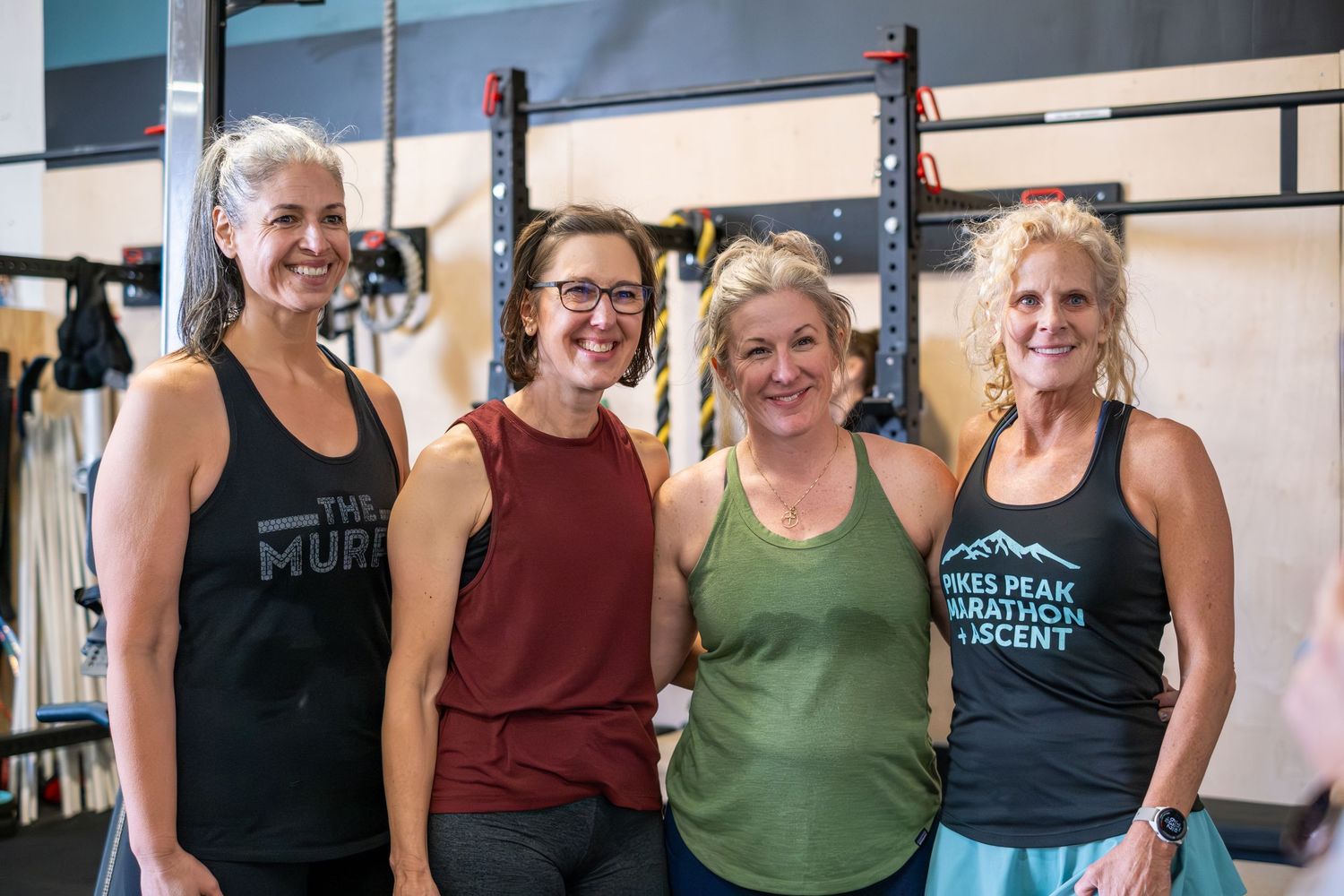 Four smiling women pose together in athletic wear inside a CrossFit gym after a workout.