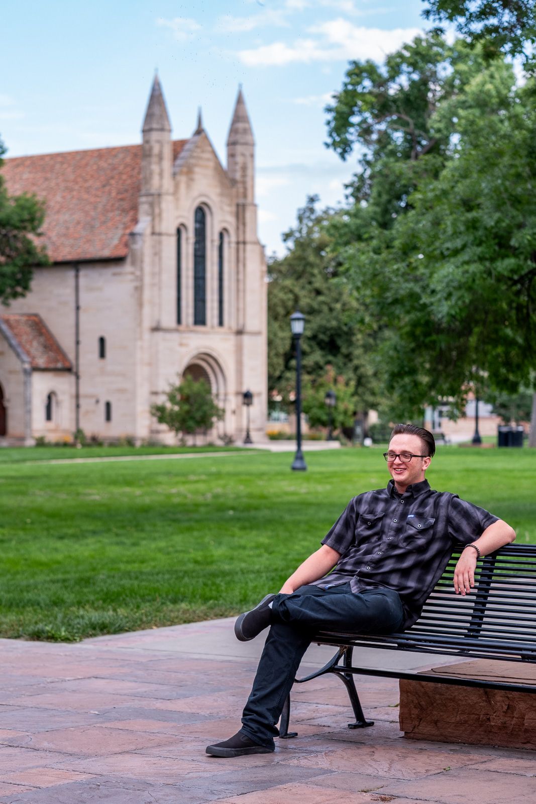 Man with glasses relaxing on a bench in front of a historic stone church on a college campus.