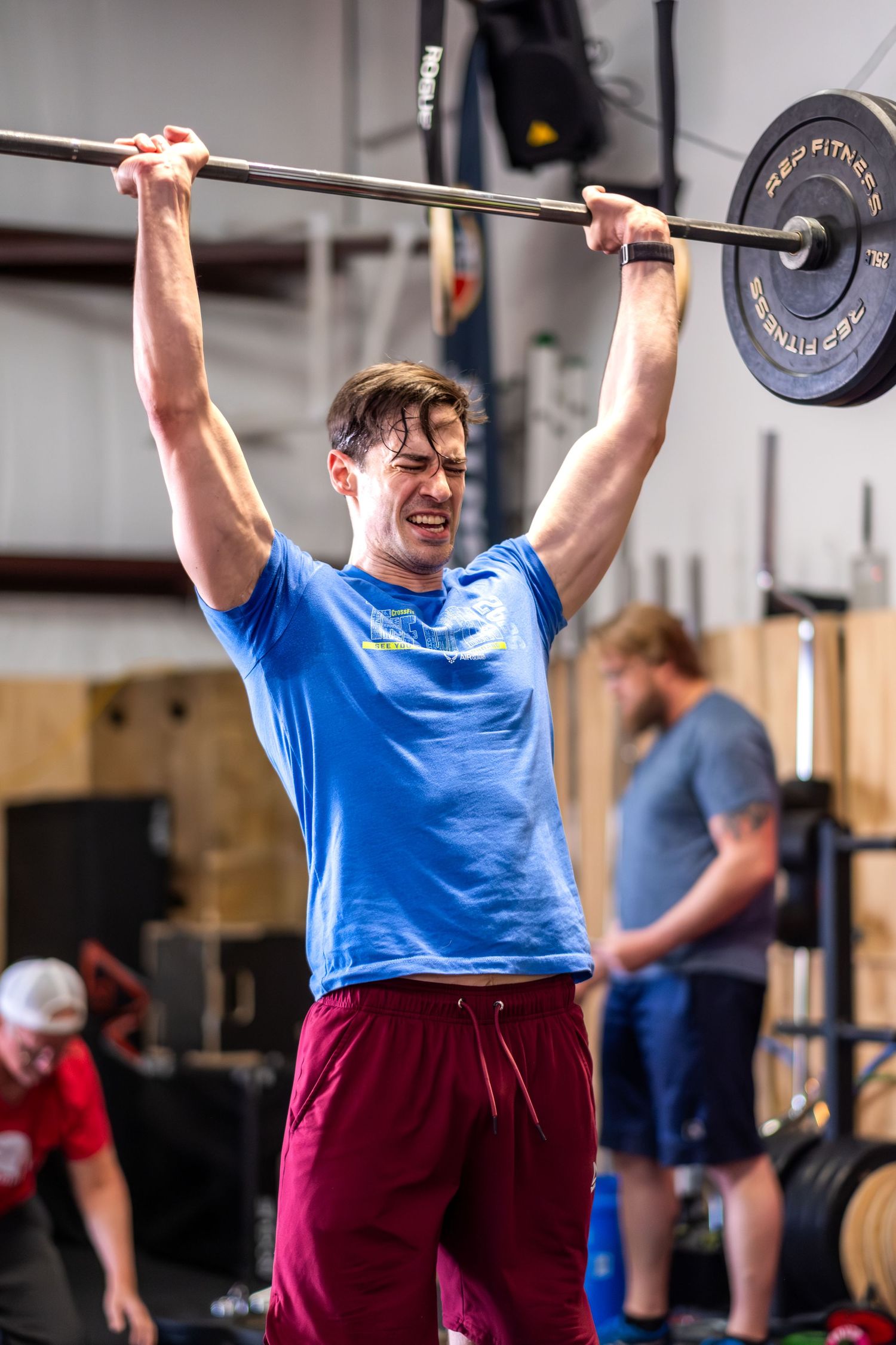 Athletic man in blue shirt pressing barbell overhead in a CrossFit gym.