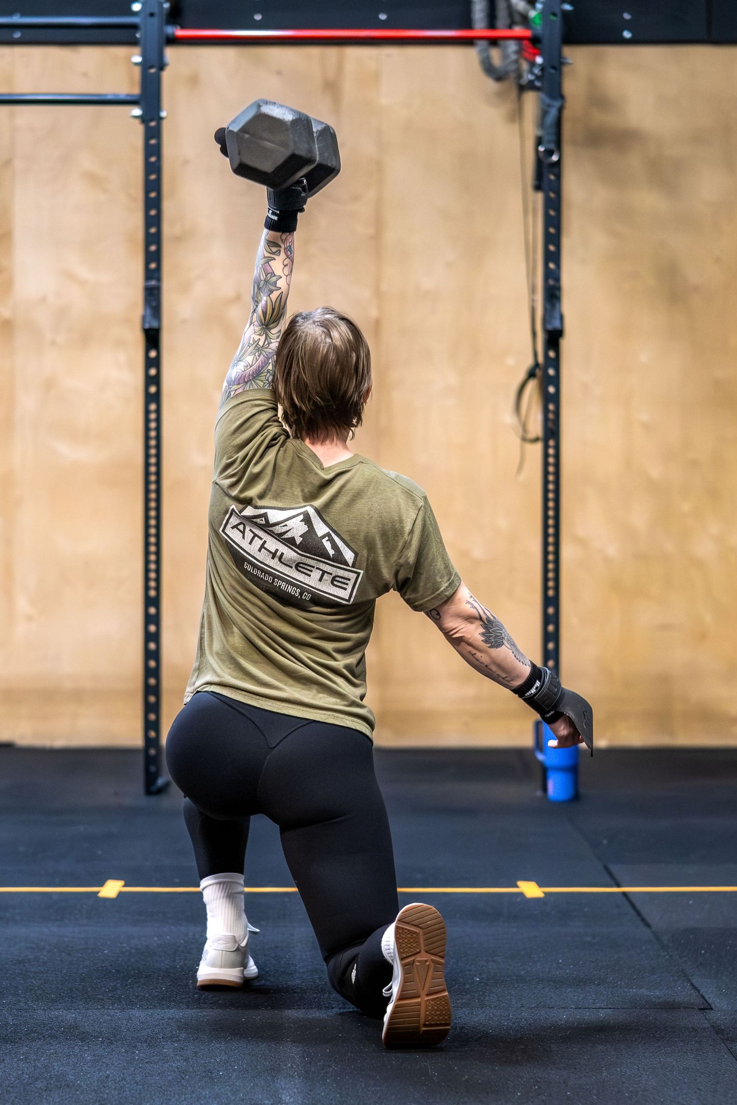 Tattooed woman in athlete shirt performs overhead dumbbell lunge in a CrossFit gym.