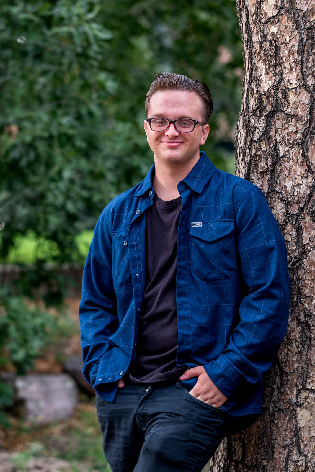 Young man in blue denim jacket leaning against a tree trunk surrounded by green foliage outdoors.