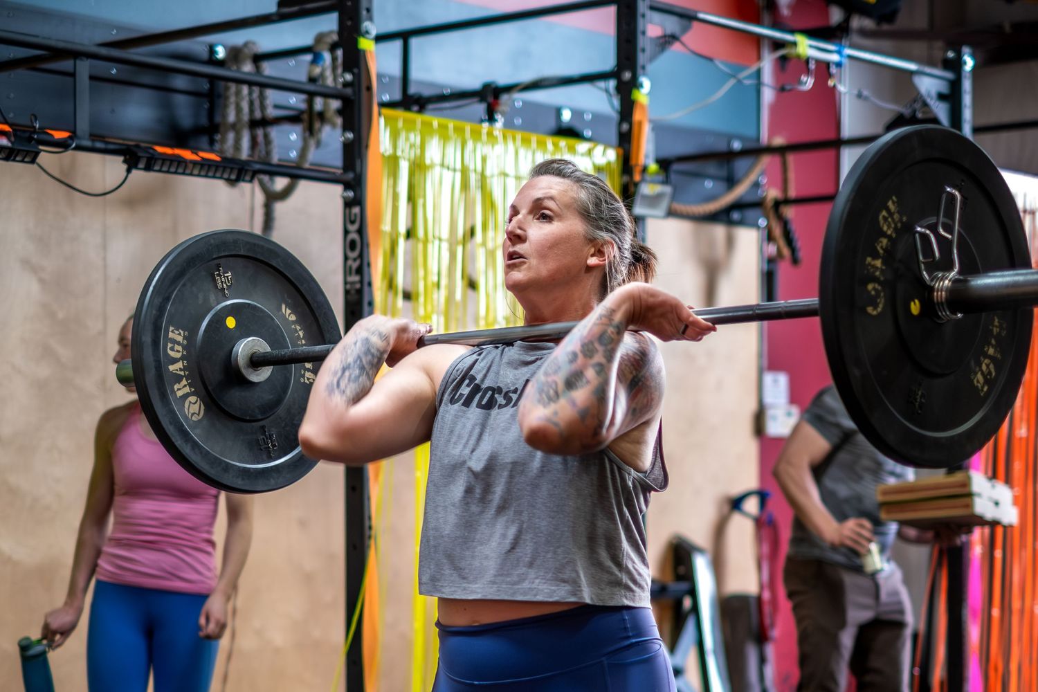 Tattooed woman performing a barbell clean lift at a CrossFit gym with weight plates.