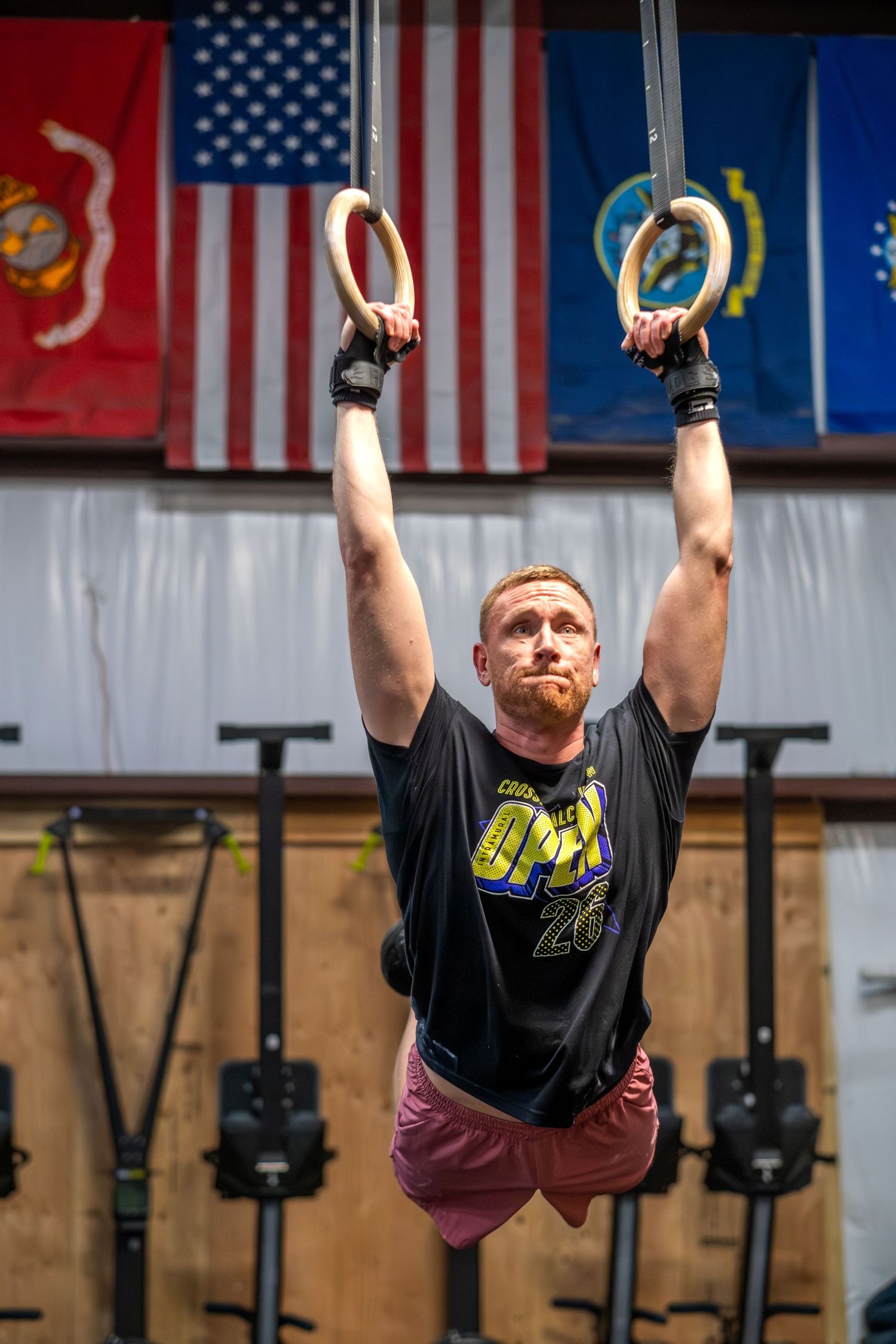 Muscular redhead man performs gymnastic ring hang exercise in CrossFit gym with flags on wall.