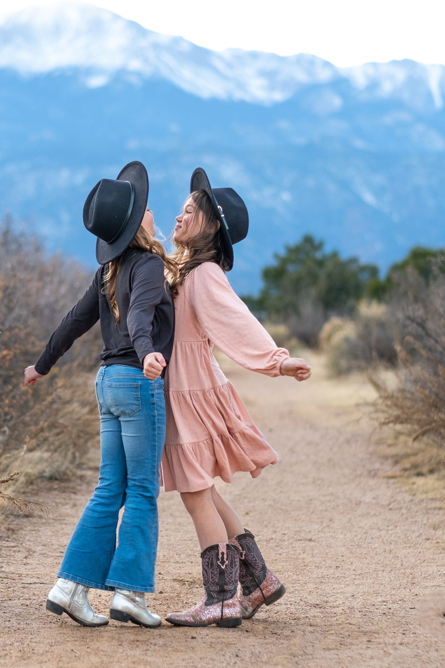 Two girls in black cowboy hats dancing on dirt path with snow-capped mountains in the background.